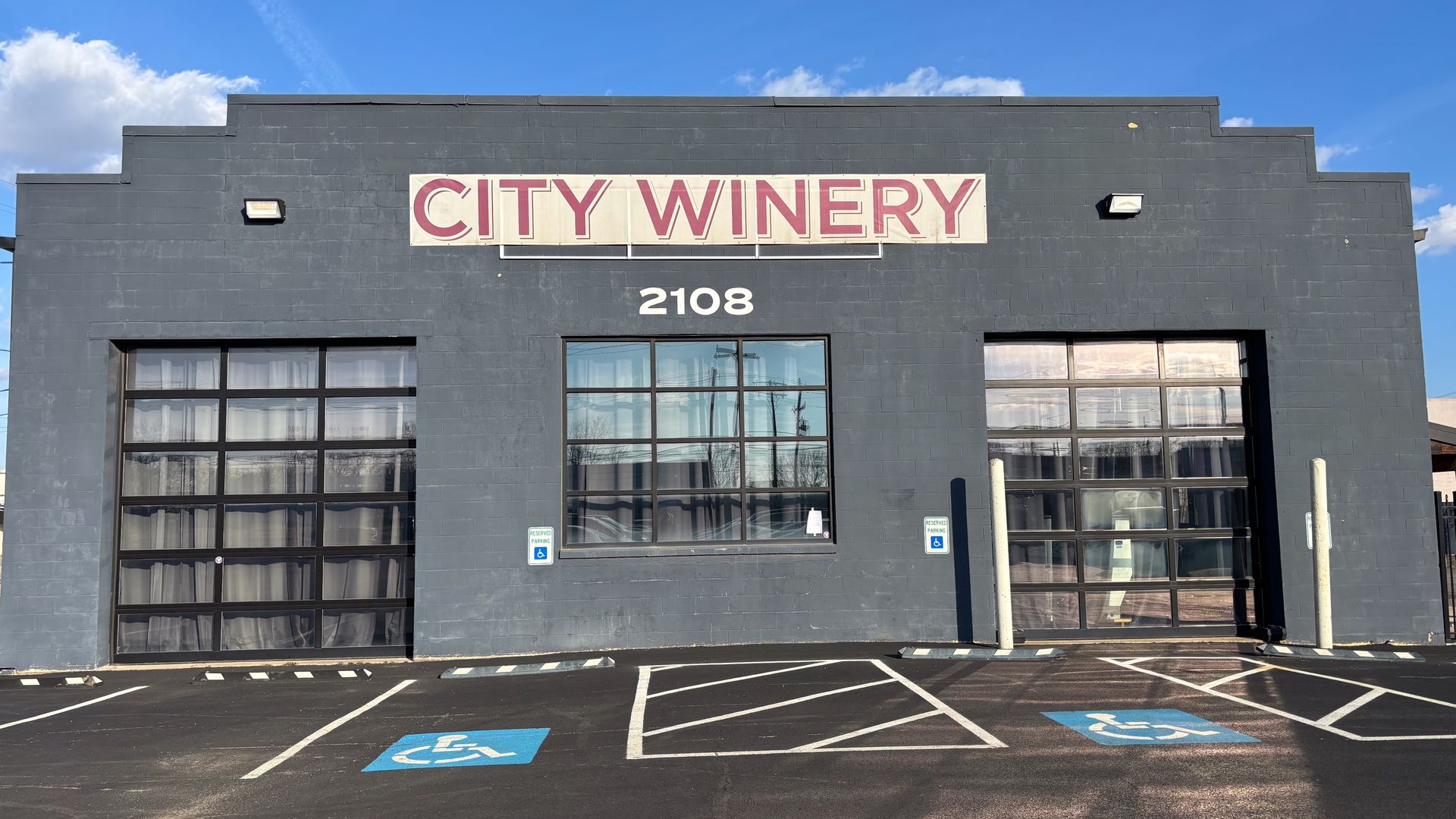 Gray building with large windows and two garage doors, labeled "CITY WINERY" with address number 2108, under a bright blue sky with a visible moon. Two handicap parking spaces in front.