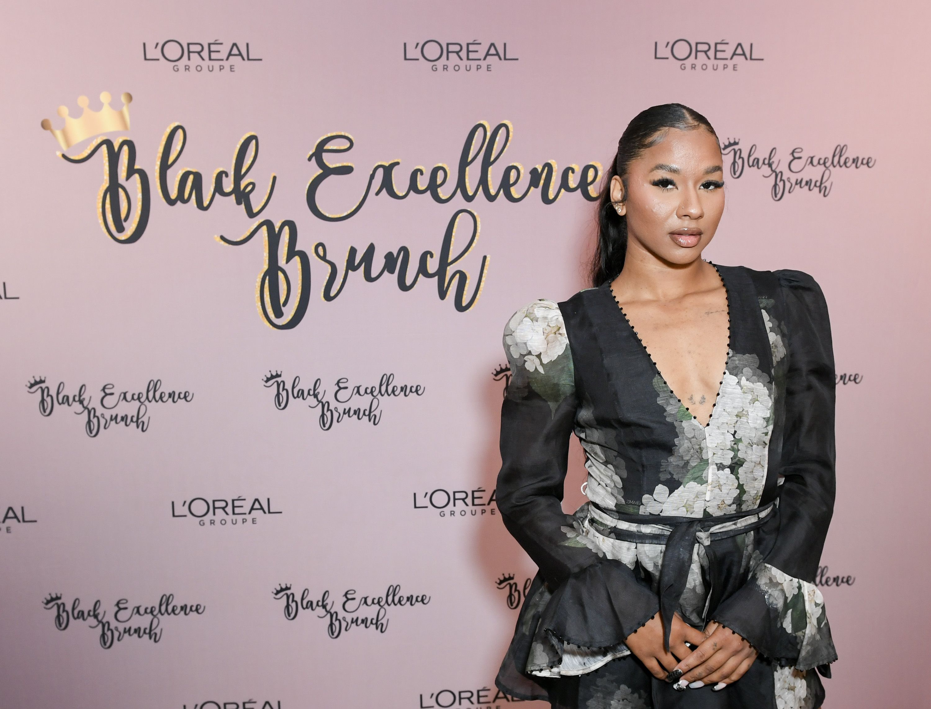 A woman in a black floral dress poses at the L'Oréal Black Excellence Brunch event with a pink backdrop featuring the event's name and L'Oréal logos.