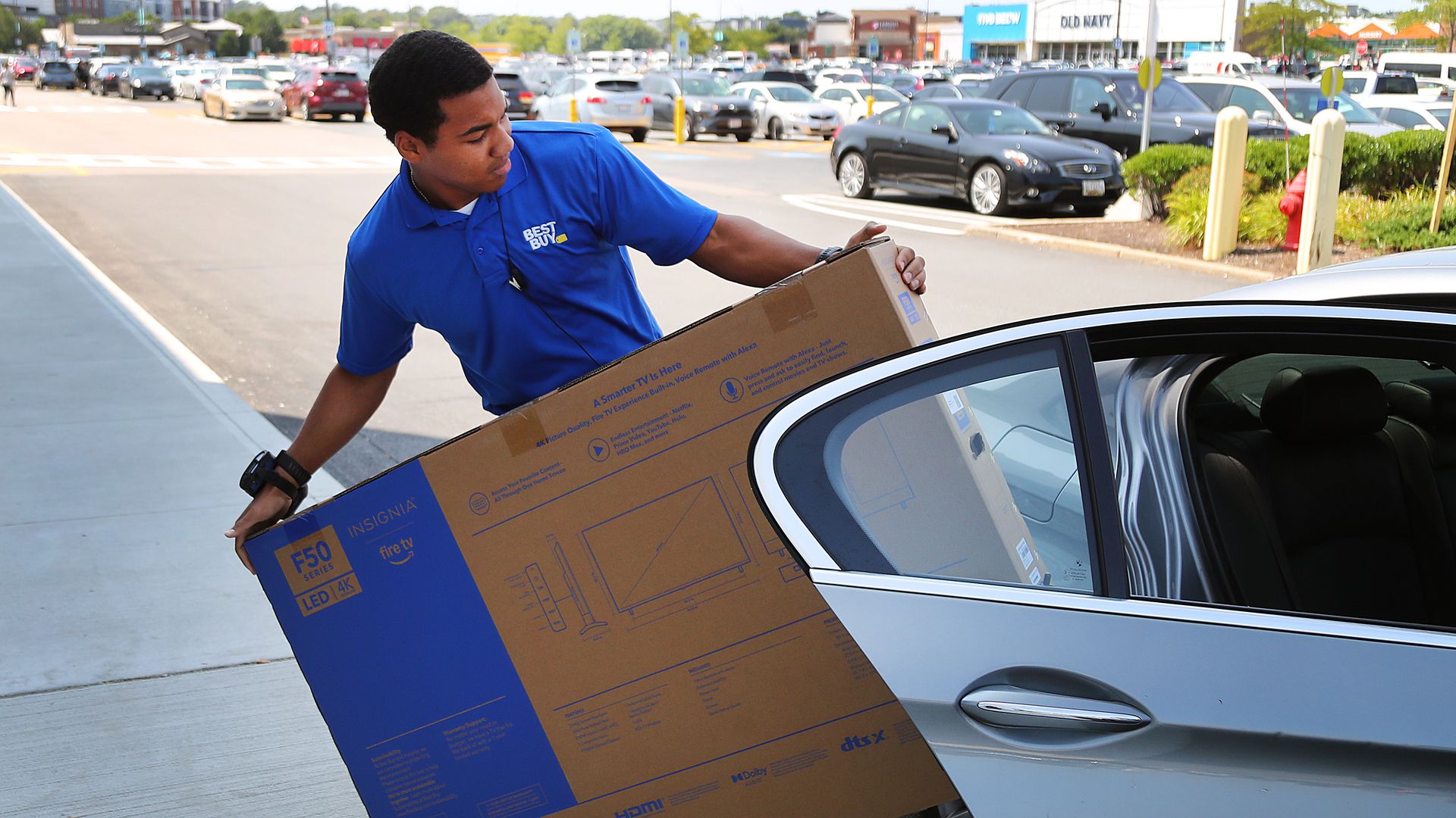 A Best Buy employee in a blue shirt loads a large Insignia F50 Series 4K LED TV box into the back seat of a silver car in a busy parking lot.