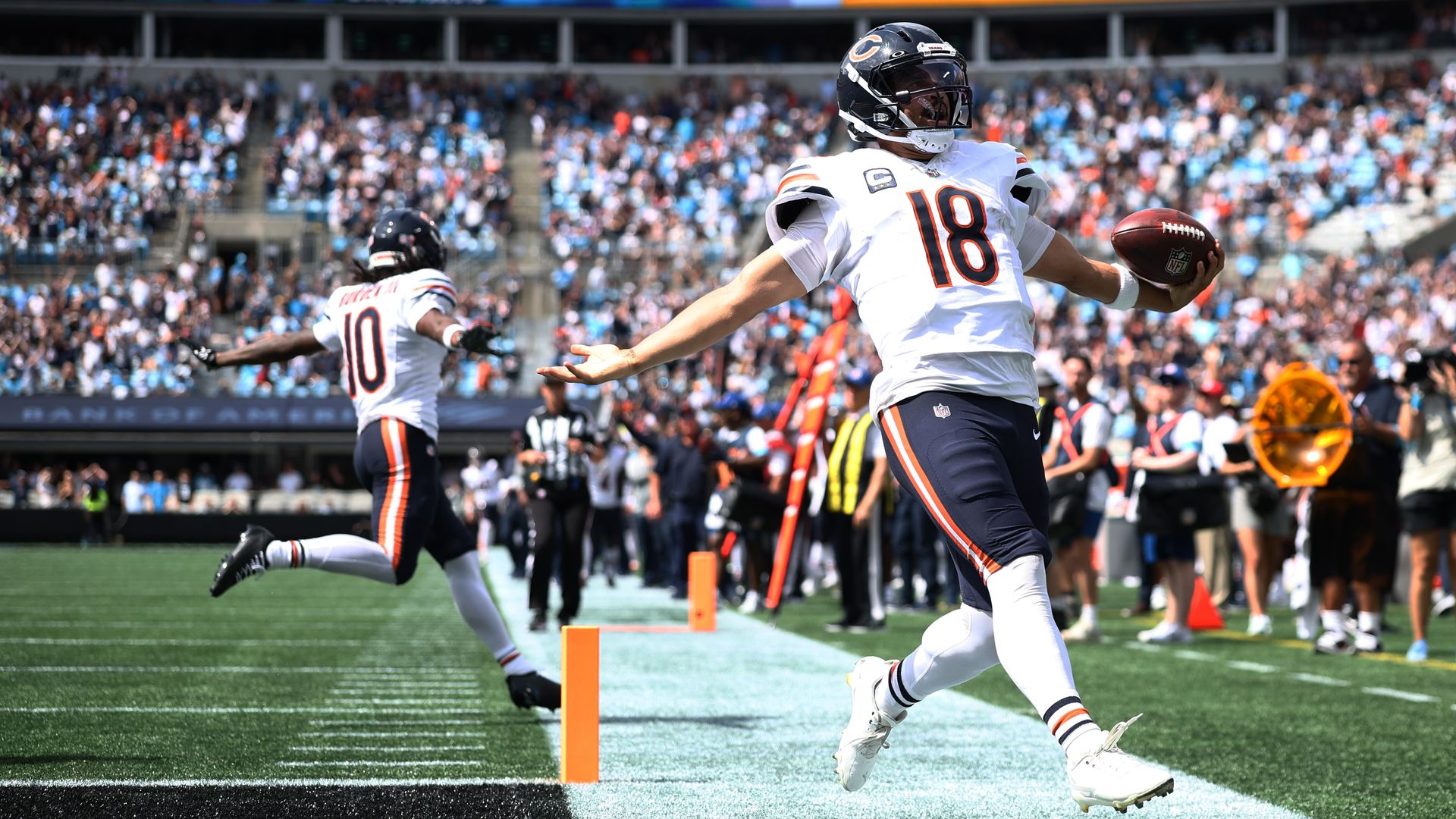 Chicago Bears player wearing #18 in a white jersey runs into the end zone, cradling a football while a crowded stadium cheers.