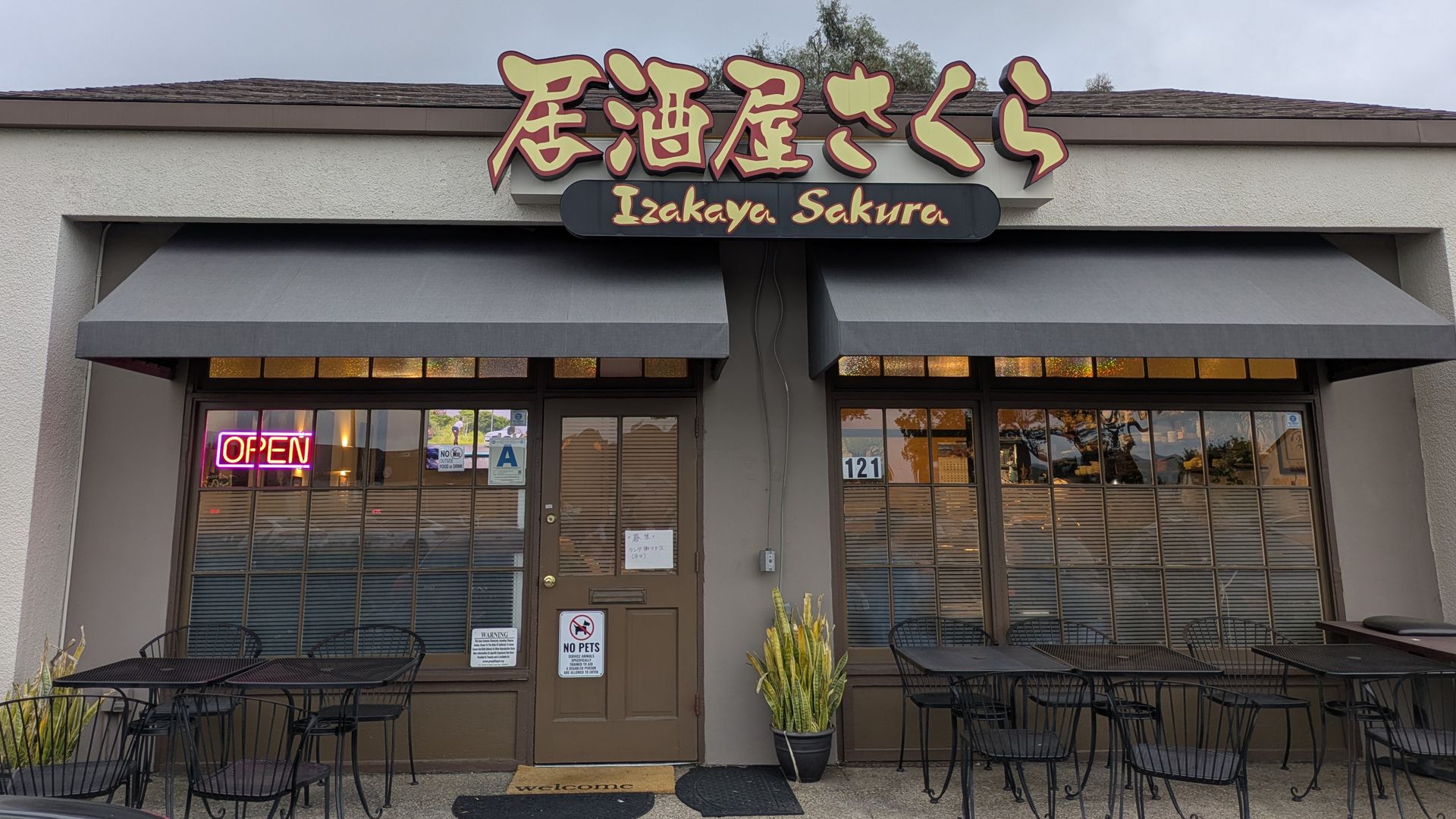Front of a Japanese restaurant with a sign in Japanese characters and "Izakaya Sakura" beneath. Neon OPEN sign in the window, gray awnings, outdoor black metal tables and chairs, and a cloudy sky.