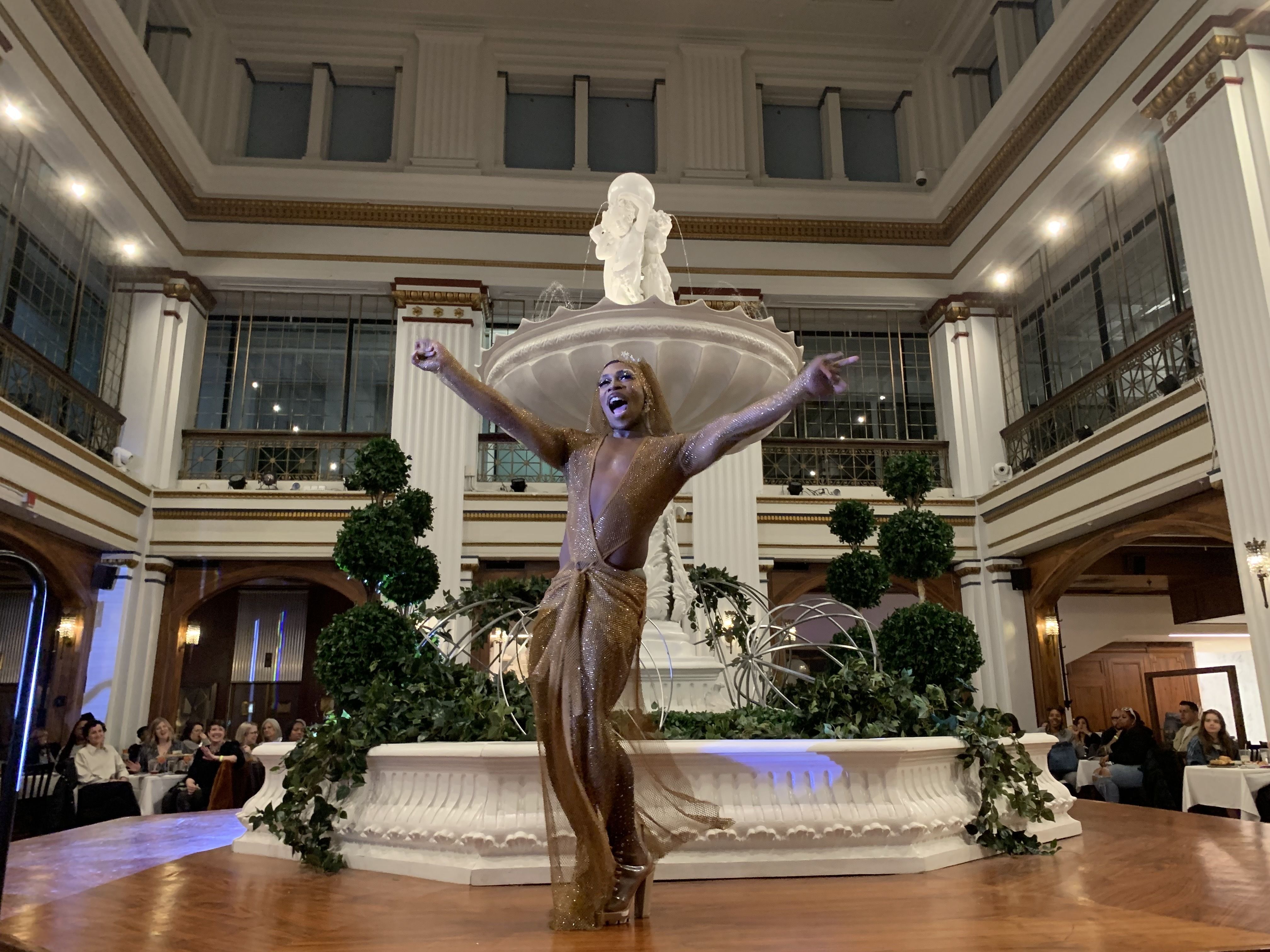 A drag performer stretches out their arms and yells to the crowd, while wearing a gold dress and high heels.
