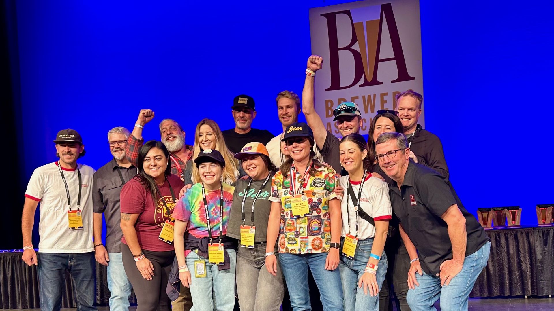 A group of 15 smiling people, most of them wearing jeans, t-shirts and yellow badges on black lanyards around their necks, smile and raise their hands in the air in a victory sign.