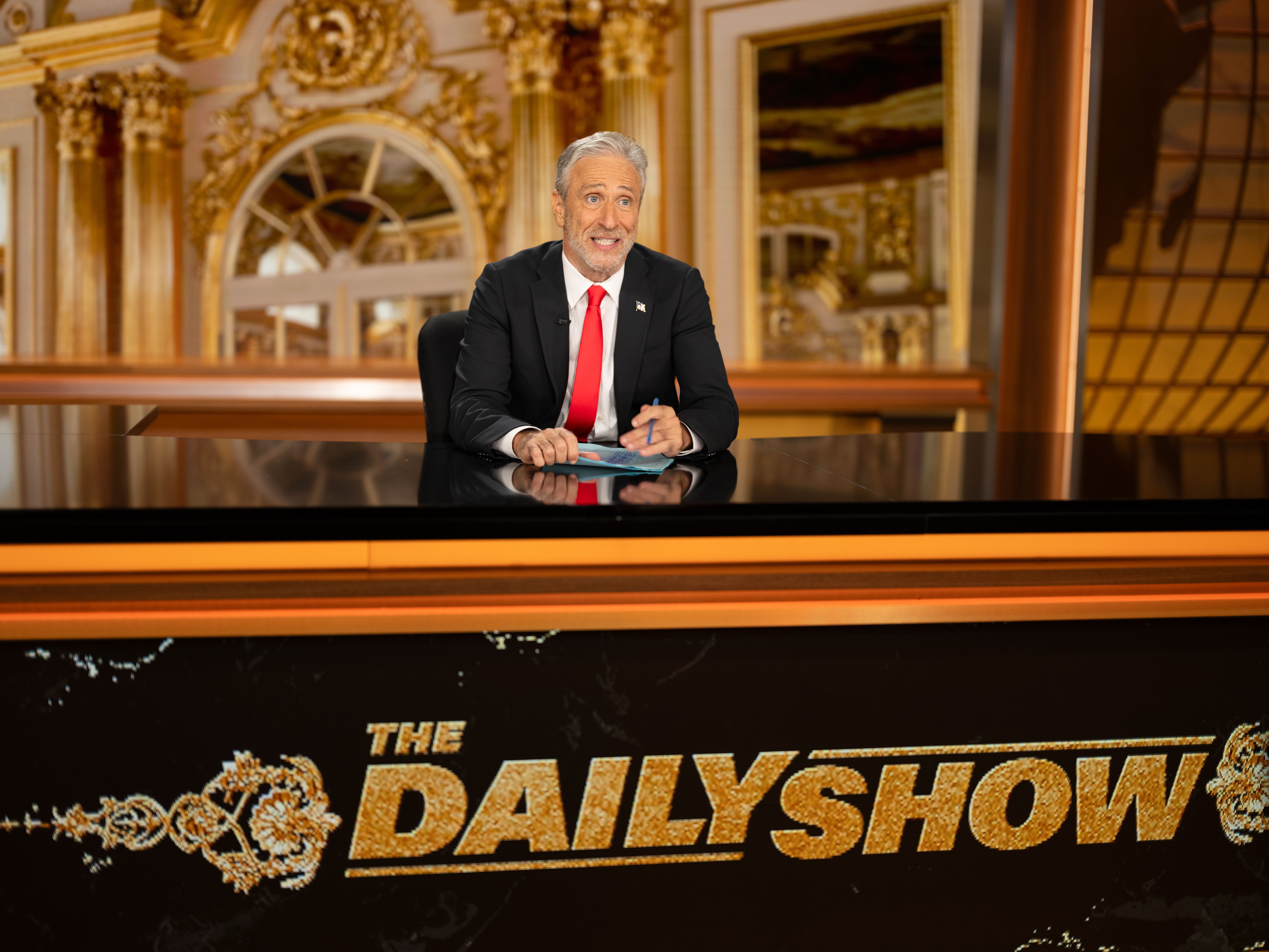 Host Jon Stewart in a black suit and red tie seated behind a desk with "The Daily Show" logo, in an ornate studio with gold decorations and large mirrors.