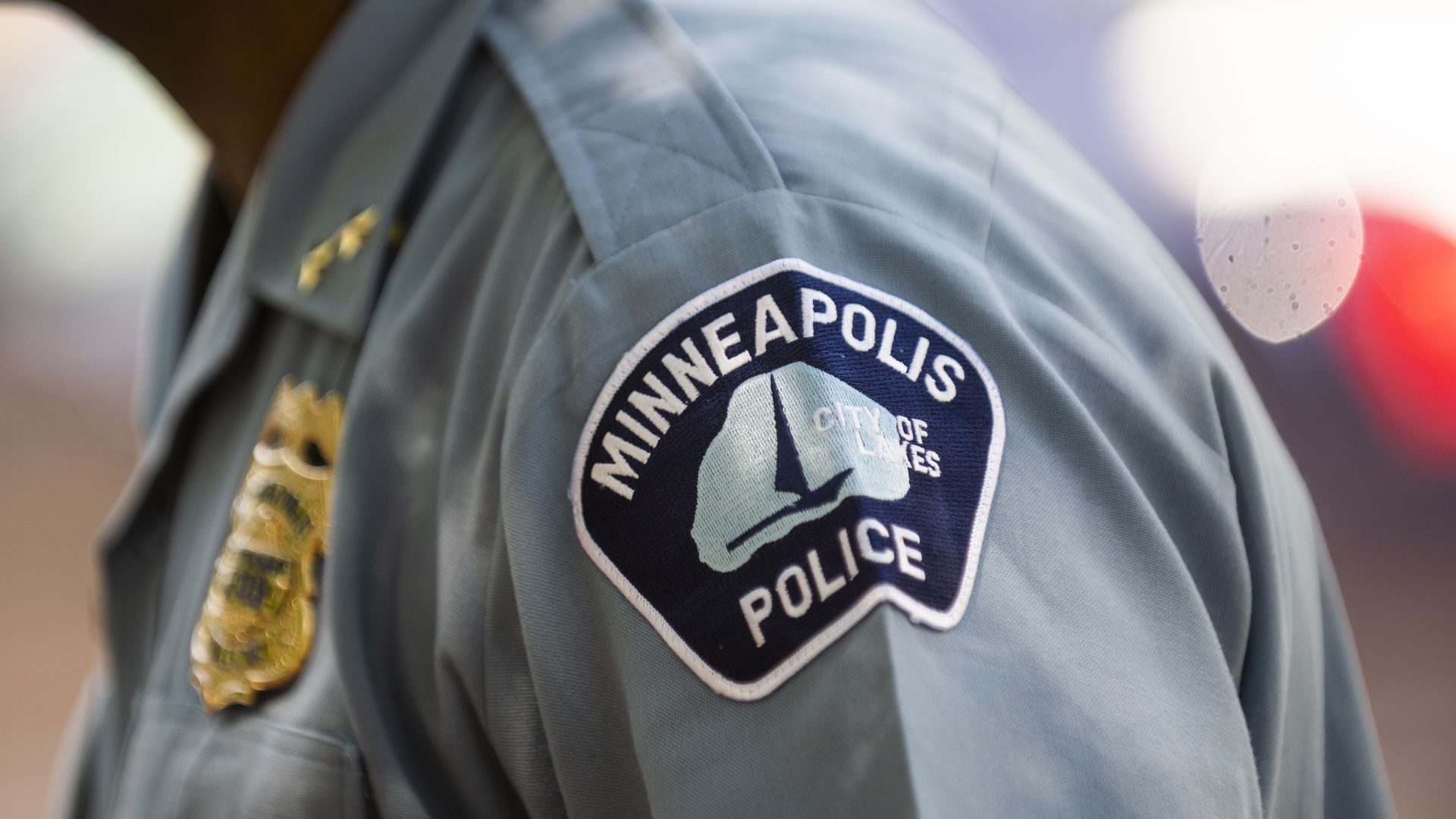 A close-up shot of a police officer wearing a powder-blue Minneapolis Police uniform. Only the officer's shoulder patch and badge are visible