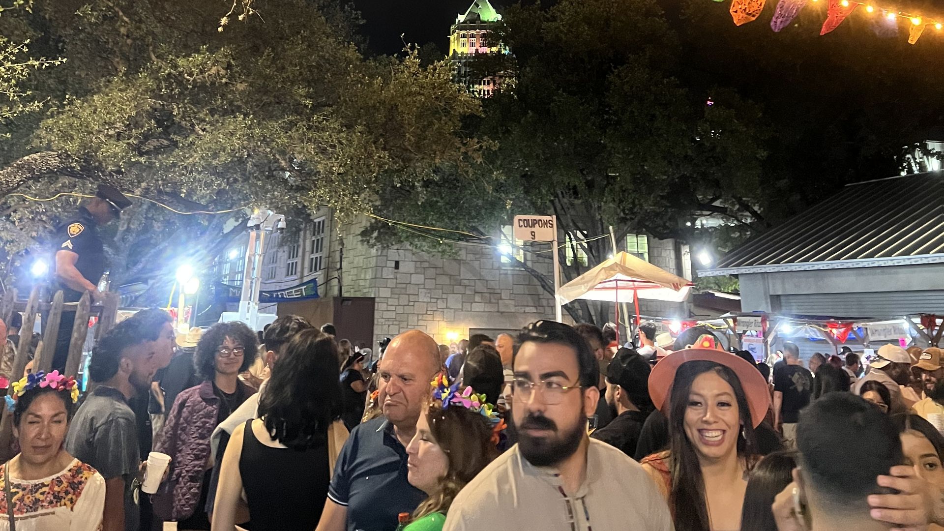 Night market under a large tree with string lights and pennants. Crowded stalls and people in the foreground, with a distant illuminated building featuring a green-domed tower against the night sky.