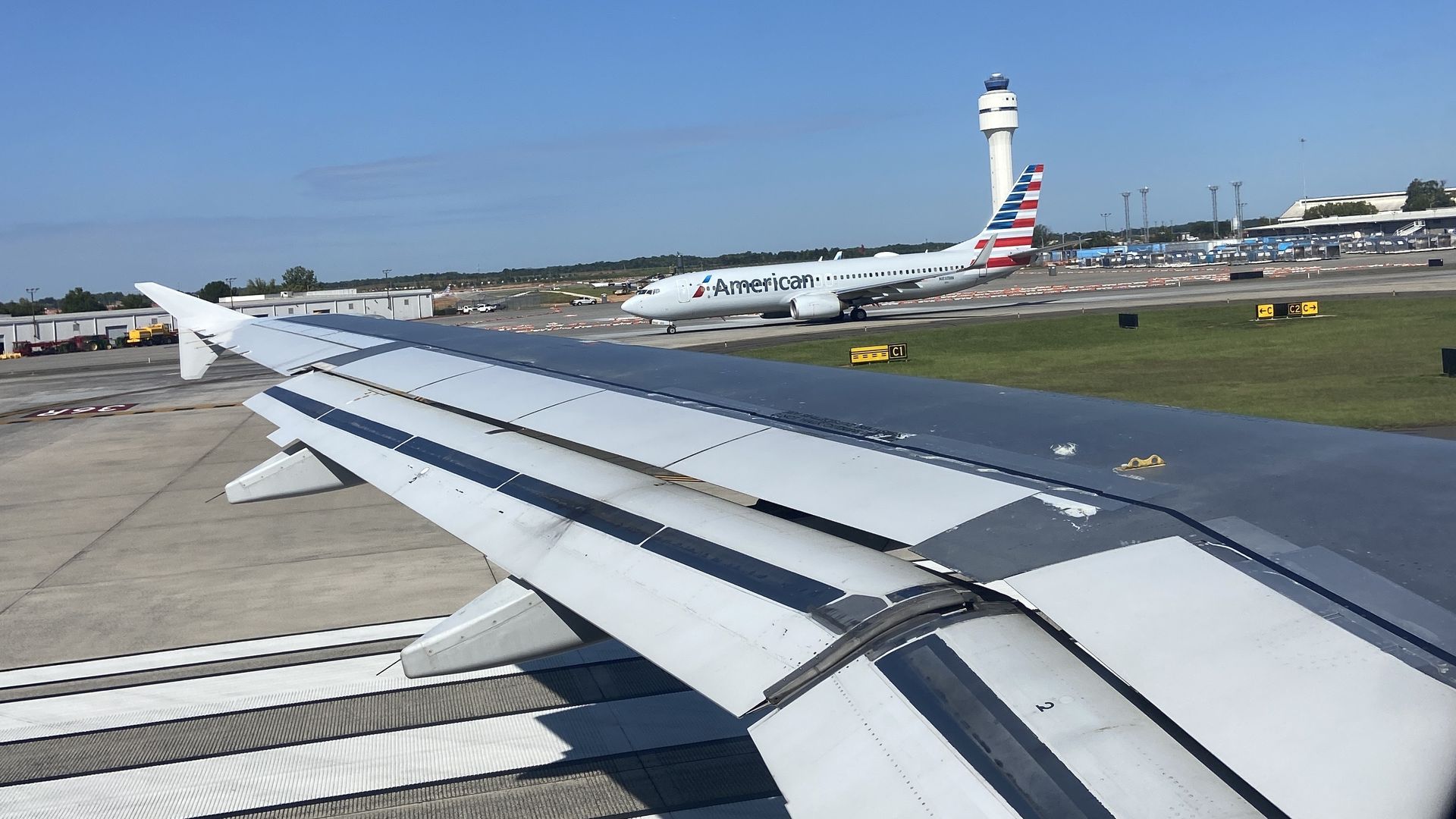 View from an airplane window: silver wing in the foreground, airport tarmac and a distant American Airlines jet, runway lights, and a tall white control tower under a clear blue sky.