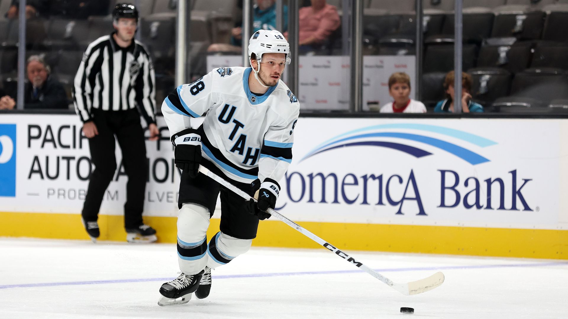 Mikhail Sergachev #98 of the Utah Hockey Club approaches the puck on the ice while a referee watches.