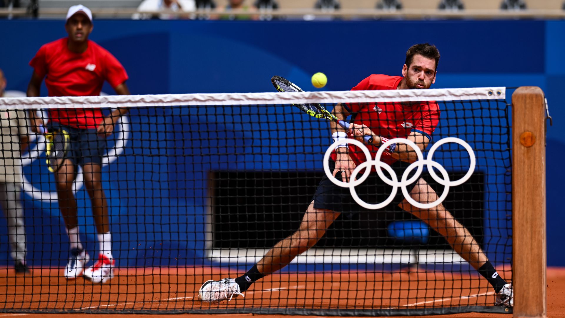 Austin Krajicek hitting the ball behind a net with the olympic rings on it