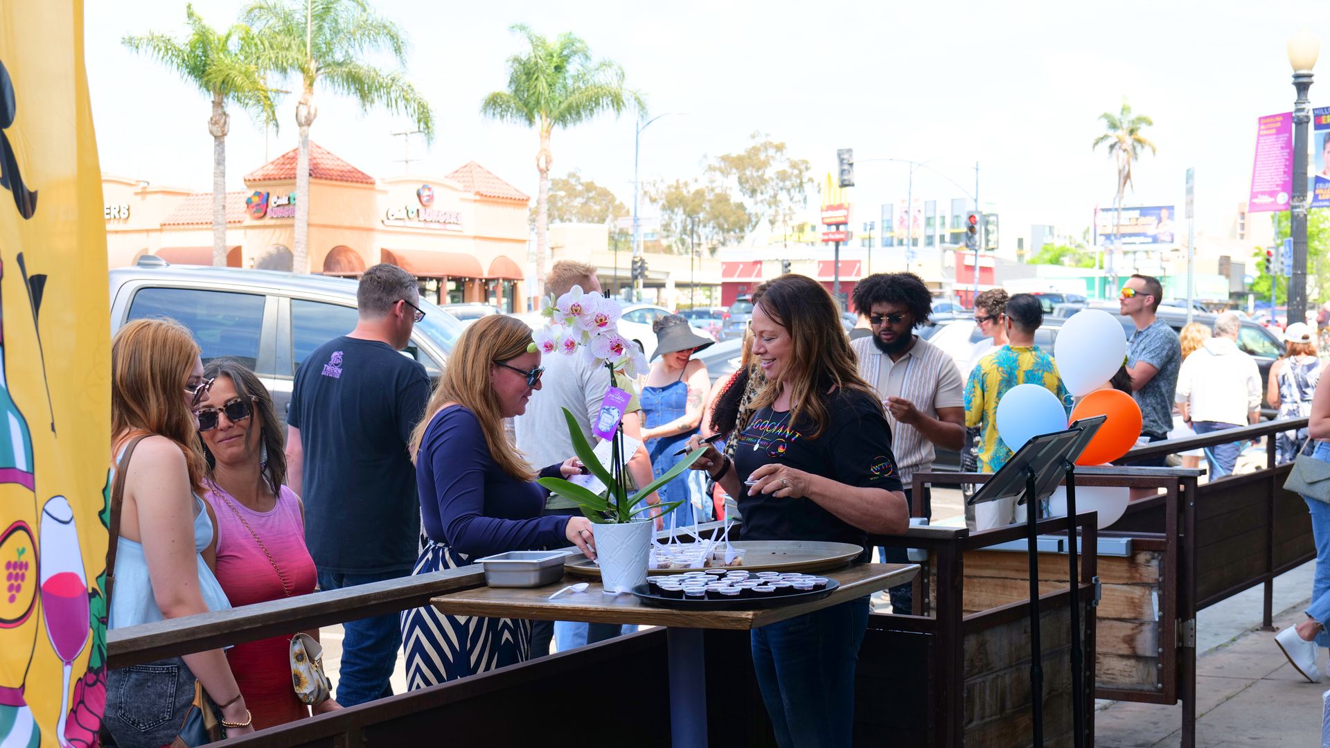 Outdoor street festival with a wooden bar; a woman in a black shirt hands small cups to a blonde woman, orchids in a pot on the table; balloons and shoppers with palm trees behind.