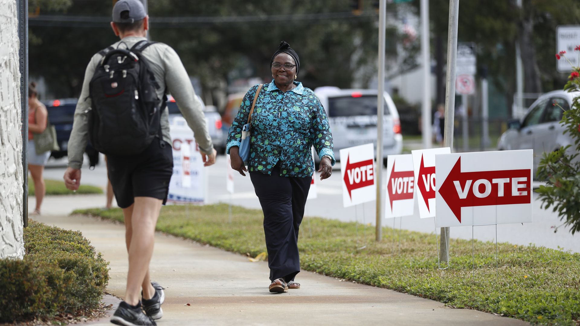 Smiling woman in teal patterned blouse and headwrap walks toward camera on sidewalk past red VOTE signs; man with backpack walks away on left; urban street with cars and traffic lights.