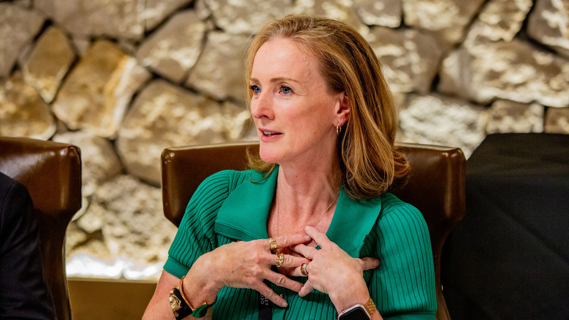 Side-profile of a blonde woman in a green blouse with gold jewelry, seated before a stone wall, hands clasped at chest, looking to the left.