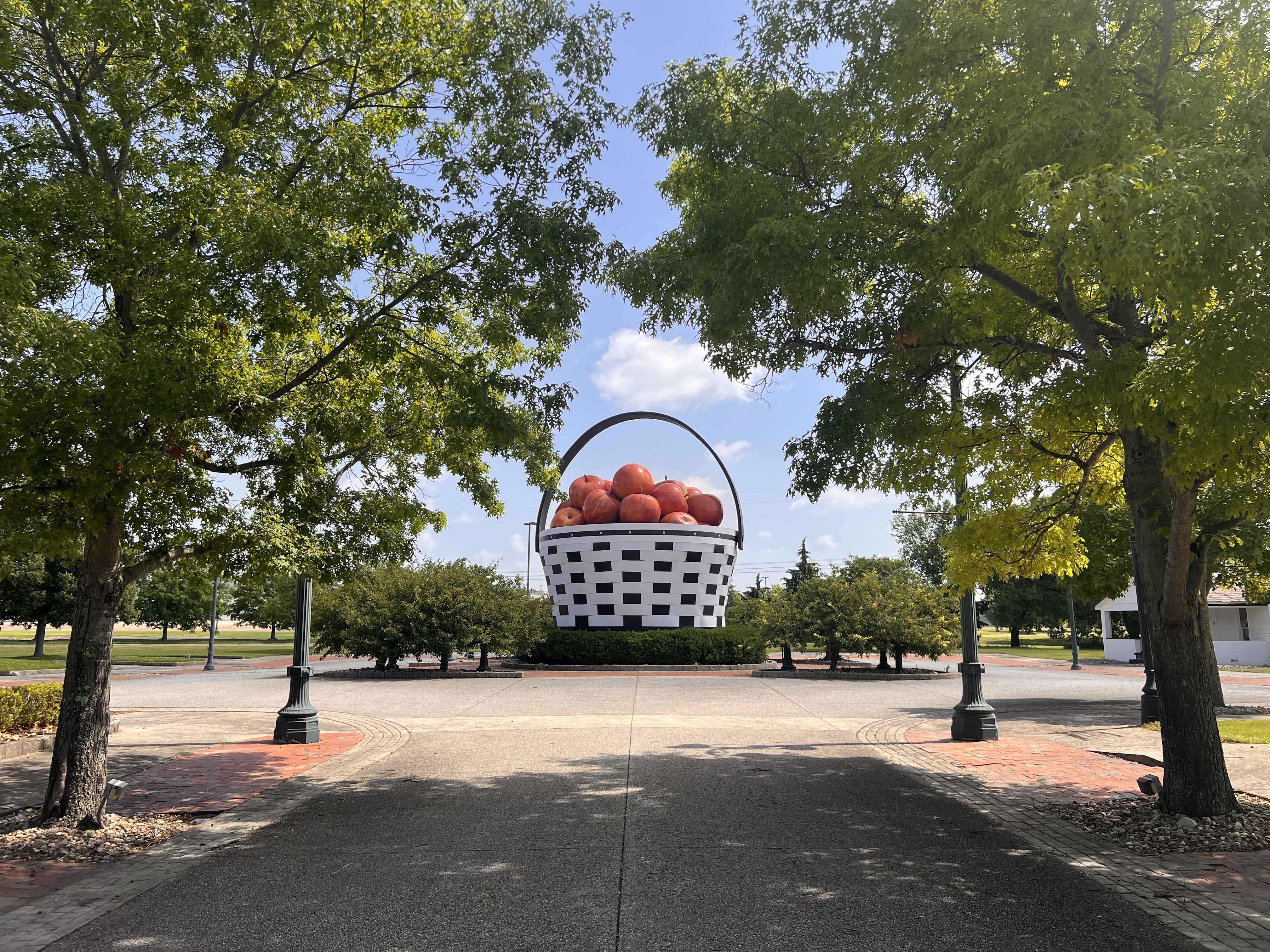 A large checkered black-and-white basket sculpture filled with red apples hangs overhead in a park, framed by green trees and a bright blue sky; lamp posts and paved walkways surround the scene.