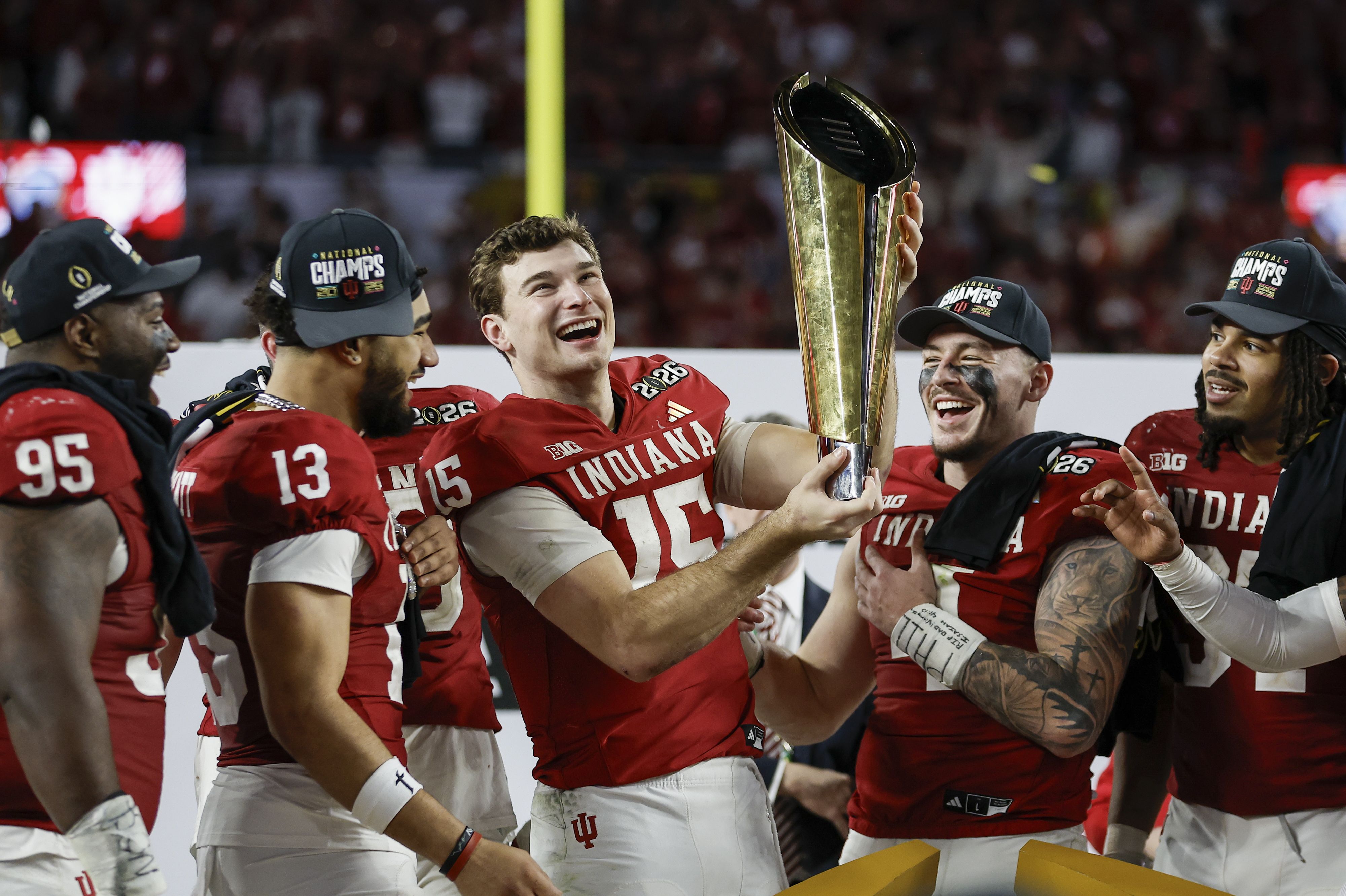 Indiana football players in red jerseys celebrating with a gold trophy, wearing black hats that say "National Champs", smiling and cheering on the field.