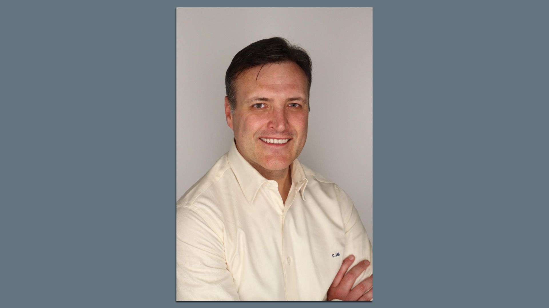 Smiling man with short dark hair wearing a light beige button-up shirt with initials C.J.M., arms crossed, against a plain light gray background.