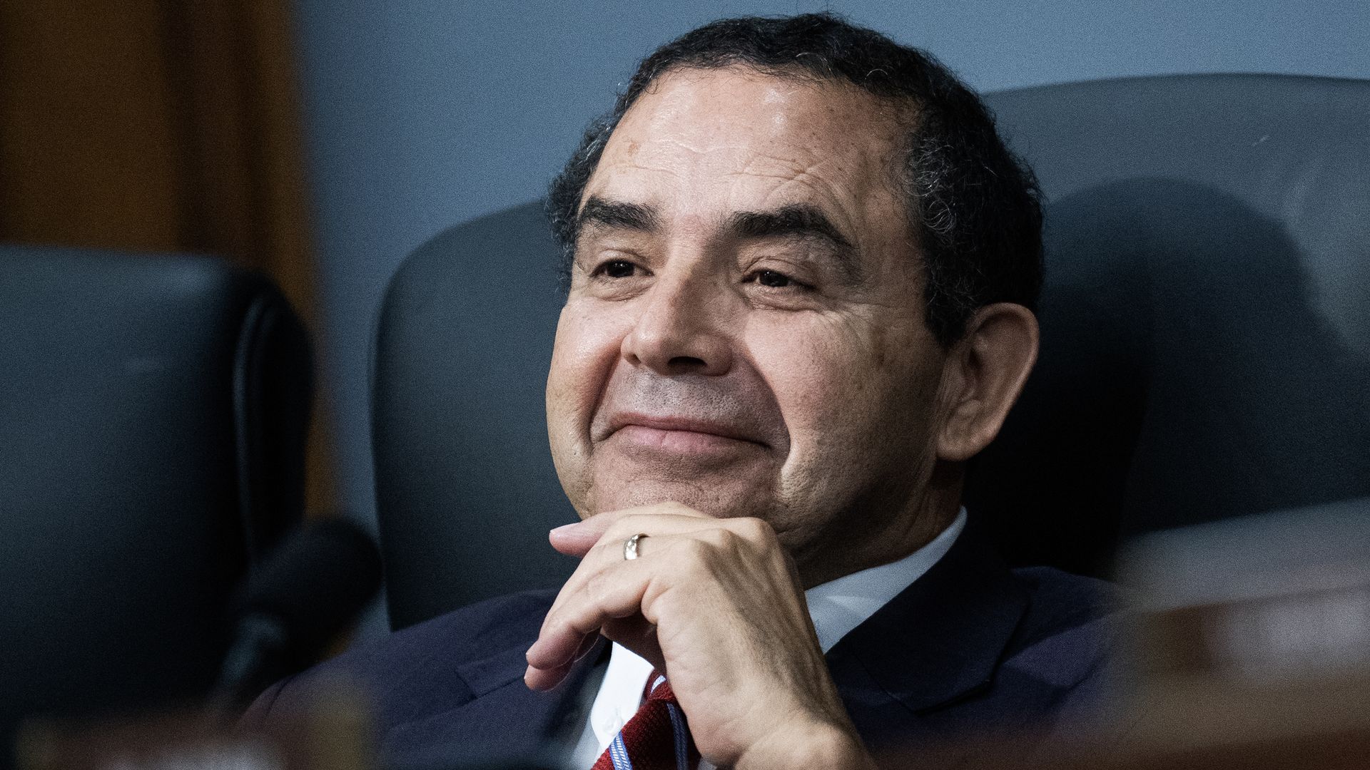 Rep. Henry Cuellar, wearing a blue suit with his hand on his chin, sitting in a black leather seat in front of a microphone.