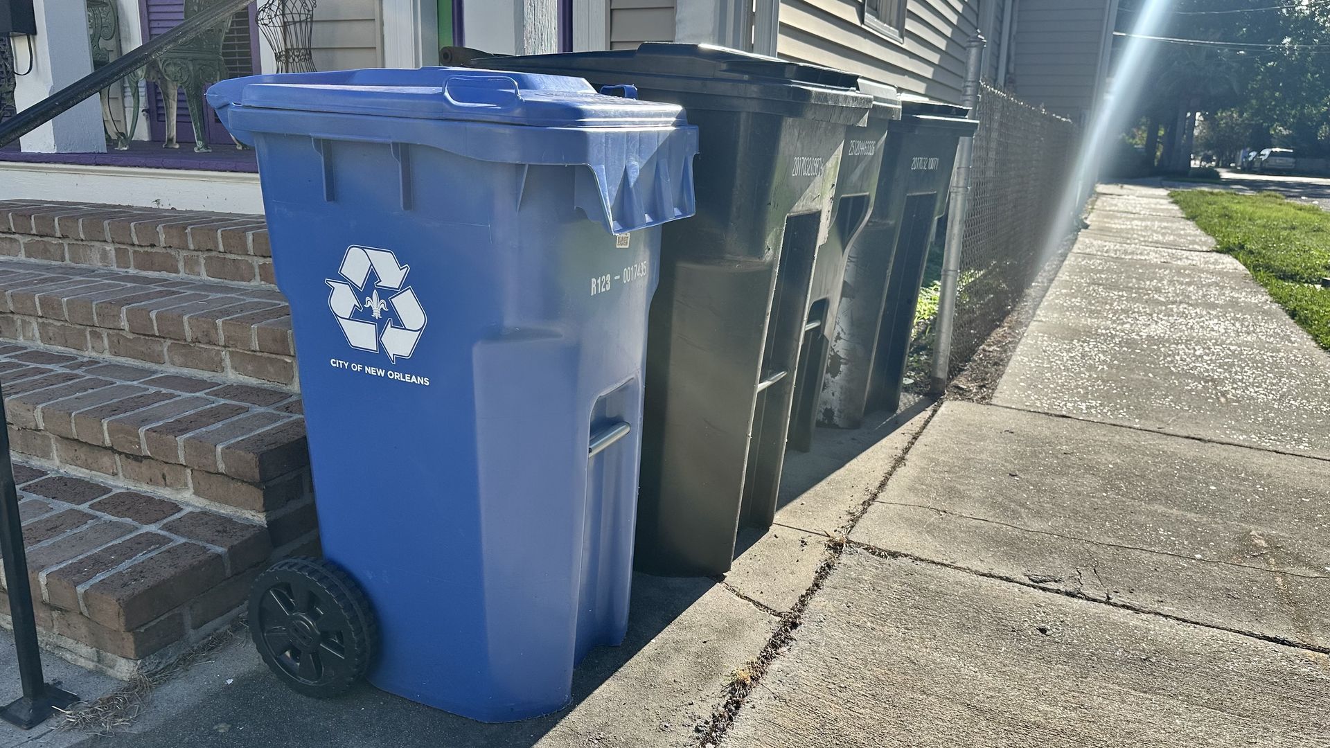 Blue recycling bin with the City of New Orleans emblem in the foreground, followed by green and black trash bins along a curb beside a brick porch, sunlit sidewalk on the right.