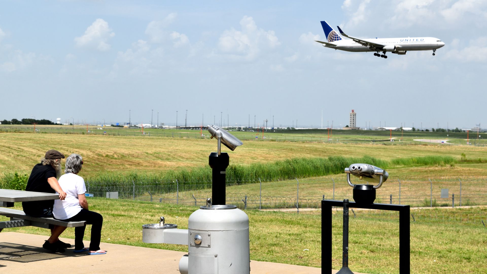 A man and woman sit on a bench and watch a plane land