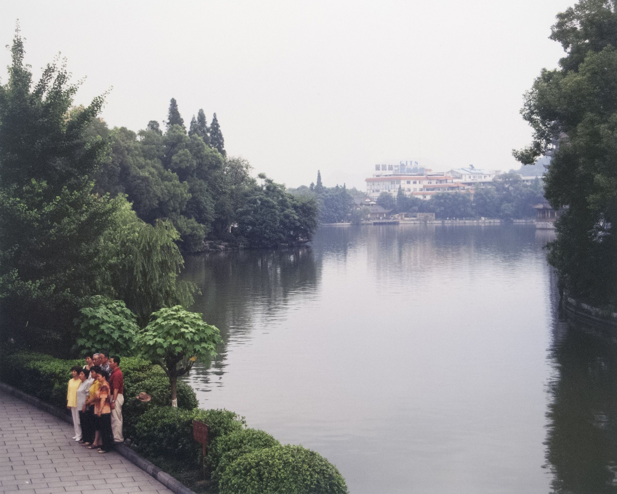 Group of people in bright clothing stand on a paved riverside path, with lush trees and a calm lake nearby; a distant white building with a red roof sits across the water.