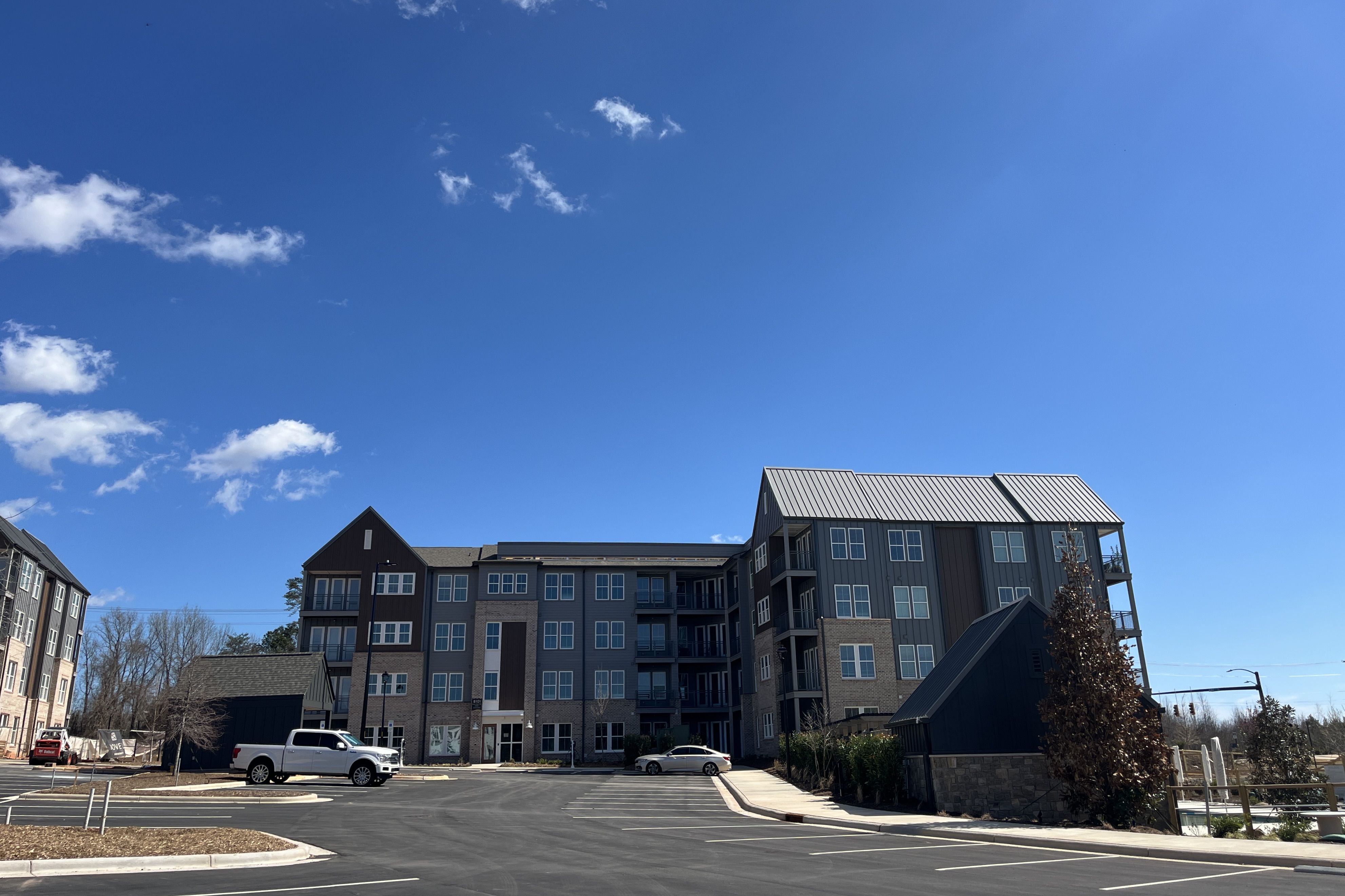 Wide view of a modern, multi-story apartment complex with gray and brown siding, balconies, and a metal roof, set against a clear blue sky; a paved parking lot with a few cars in the foreground.