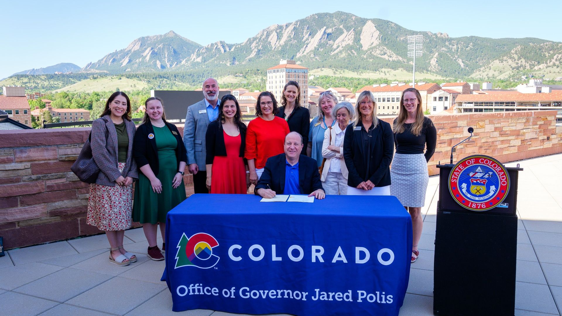 Group of people posing outdoors behind a blue Colorado banner at a podium, with the seal and text "Office of Governor Jared Polis", against a scenic mountain backdrop.