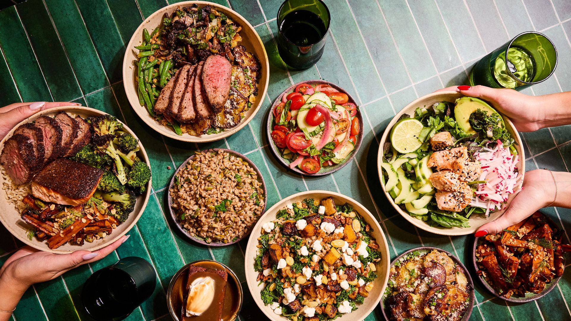 Top-down view of a green tiled table with various bowls of colorful food including sliced steak, salmon, roasted vegetables, grain salad, fresh cucumber and tomato salad, mixed greens with avocado and grilled salmon, and a dessert with cream.