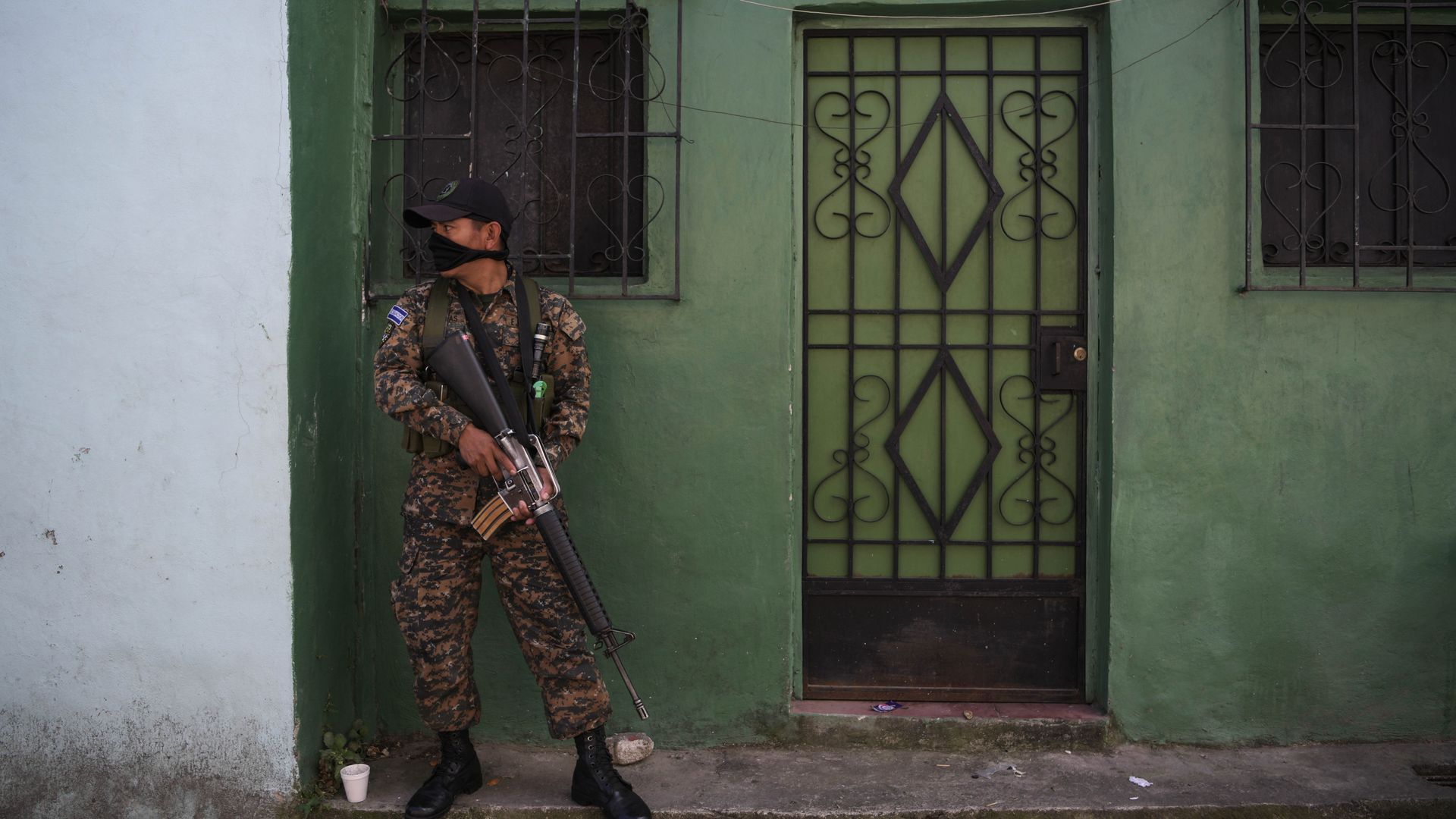 In this image, a Salvadoran army soldier patrols in a neighborhood dominated by the MS-13 gang in San Salvador. The soldier stands against a green wall, carrying a rifle.