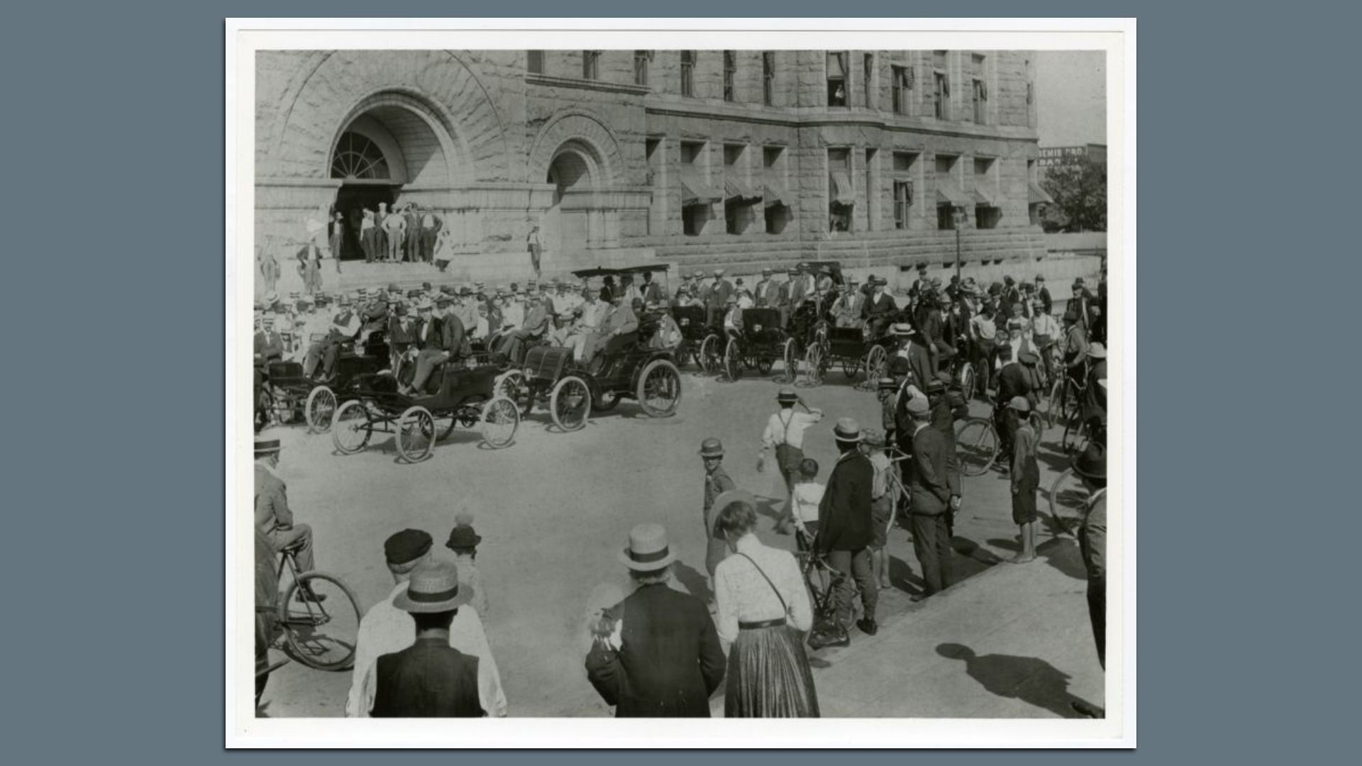 Black-and-white photo of an early 1900s street scene with vintage cars and a crowd of men, women, and children in hats and period clothing outside a large stone building.