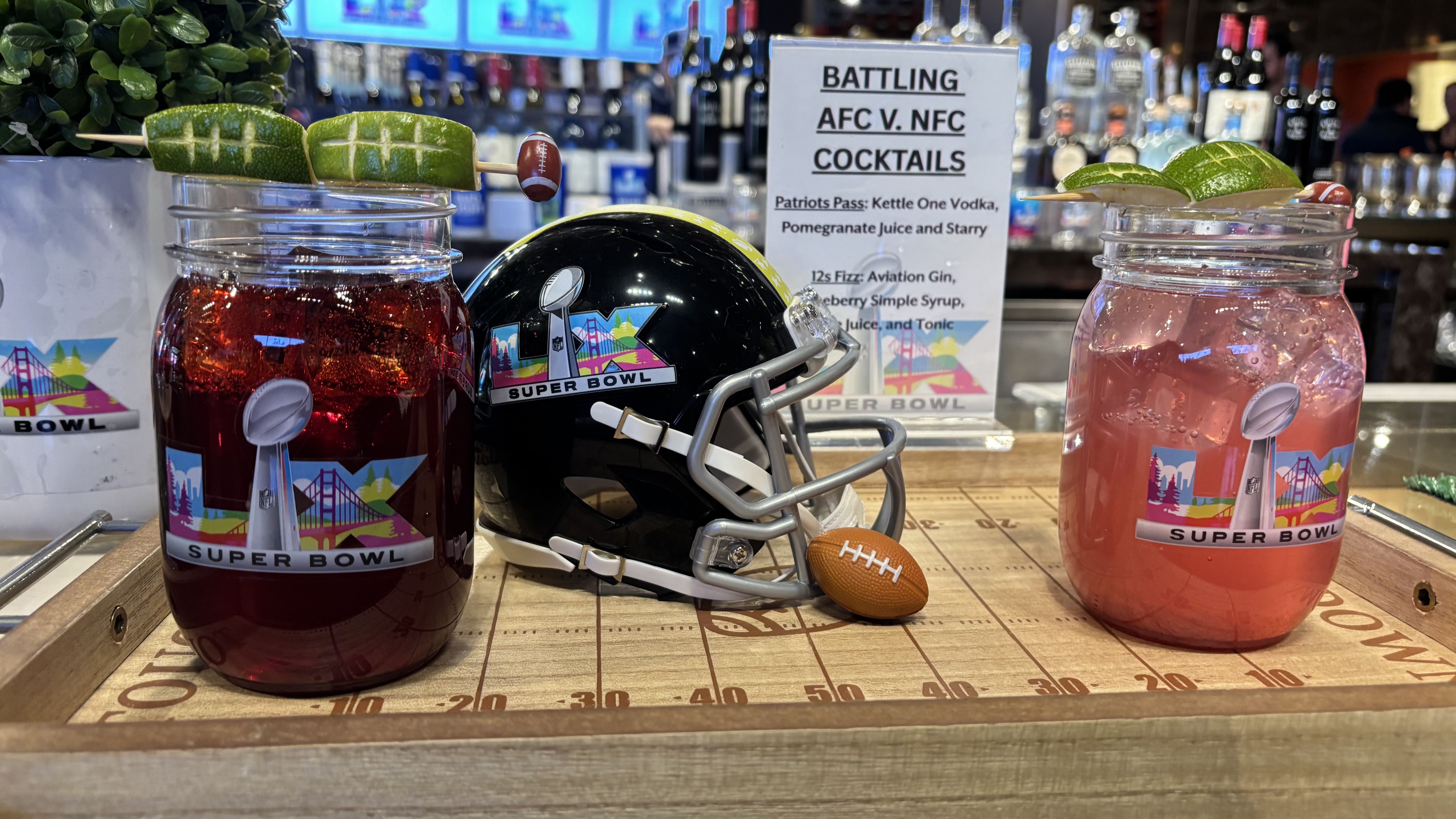 Two Super Bowl-themed cocktails in mason jars with lime garnishes, a mini black Super Bowl LIX helmet, and a sign listing AFC vs NFC cocktail ingredients on a wooden football field tray.