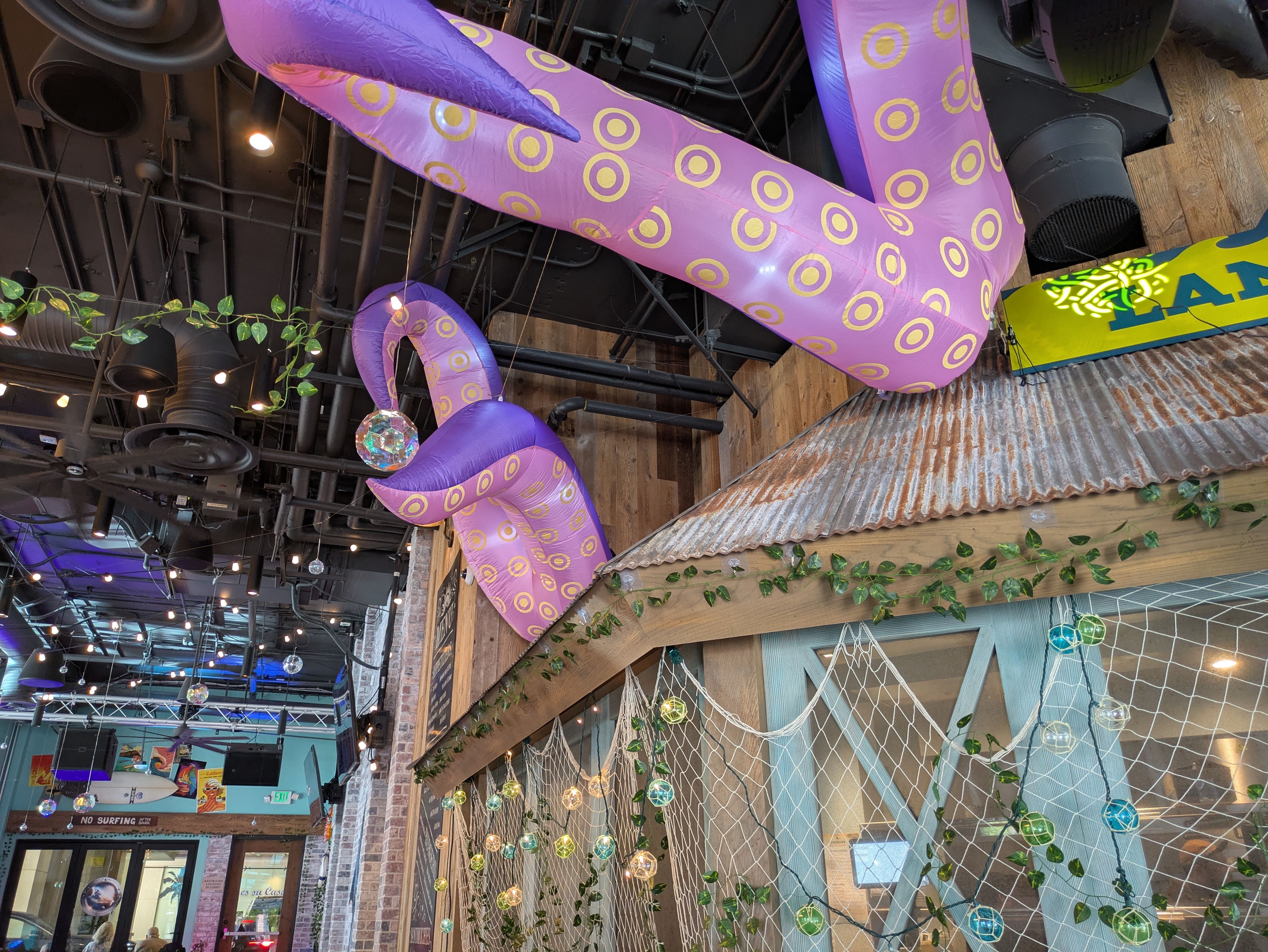 Indoor scene with large purple inflatable tentacles patterned with yellow rings draped across a dark ceiling. String lights, green vines, wooden beams, and a yellow-blue sign visible.