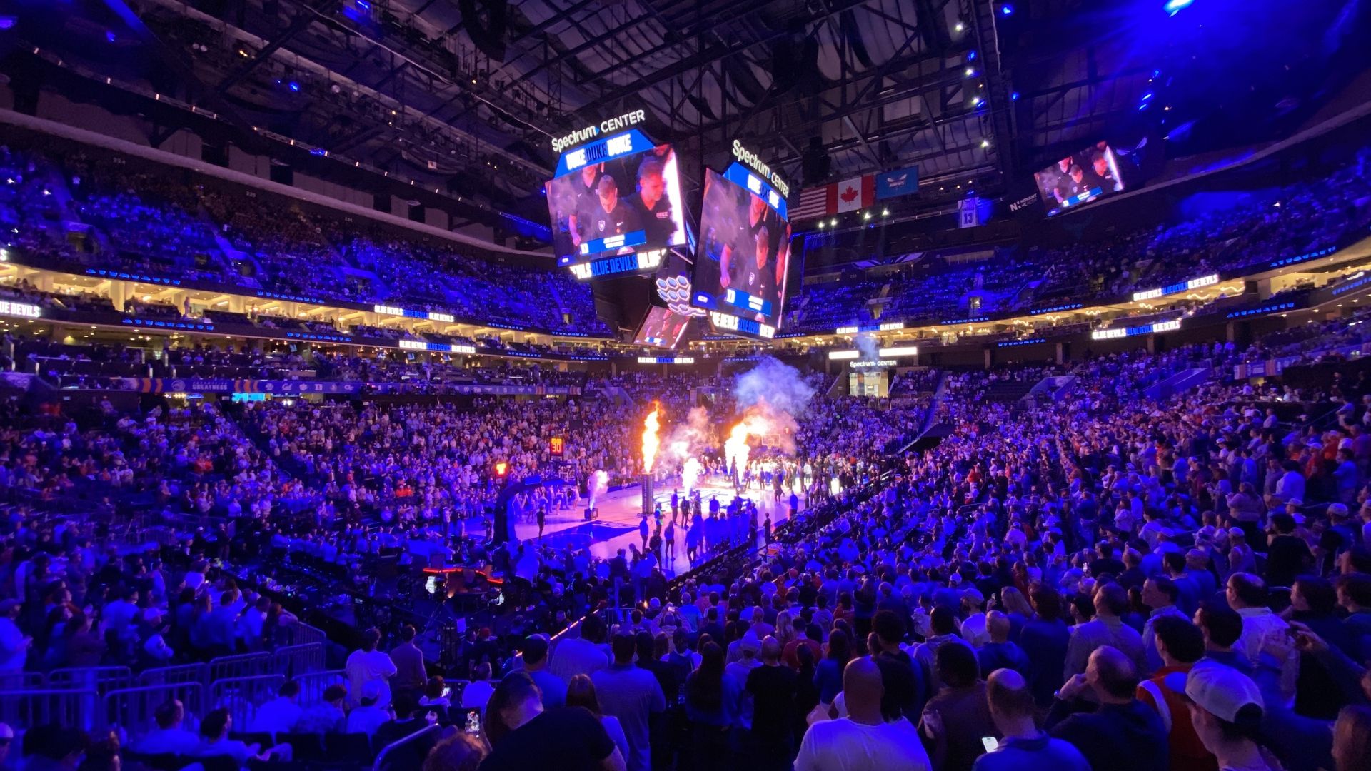 Packed Spectrum Center arena with blue lighting and large screens showing "Duke Blue Devils" before a basketball game with fire and smoke effects on the court.