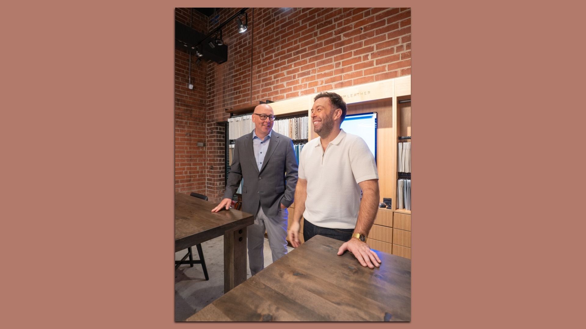 Two men smile in a brick-walled showroom with fabric swatches on a wooden display behind them; one in a gray suit and glasses, the other in a white polo shirt, standing beside dark wooden tables.