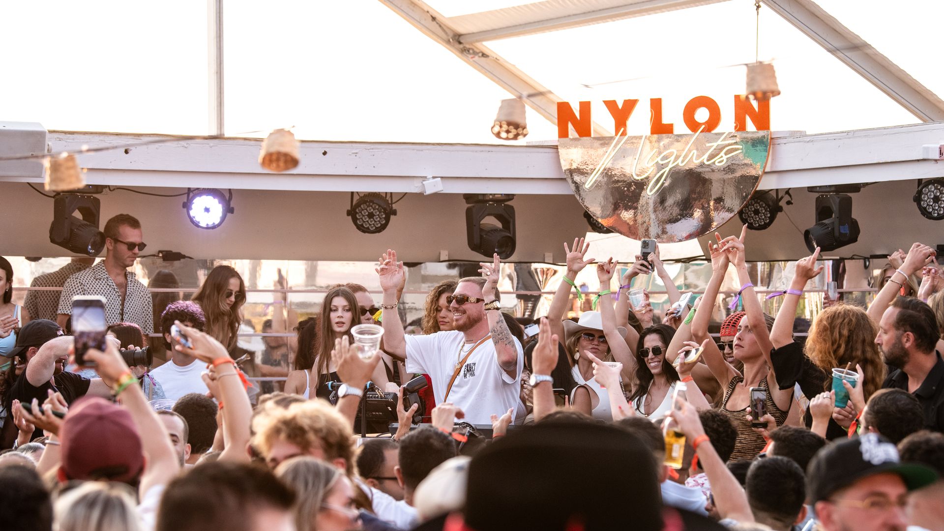 Crowd of people dancing and raising hands at an outdoor party under a tent with a sign reading "NYLON Nights," during daylight with festive atmosphere and mixed casual clothing.