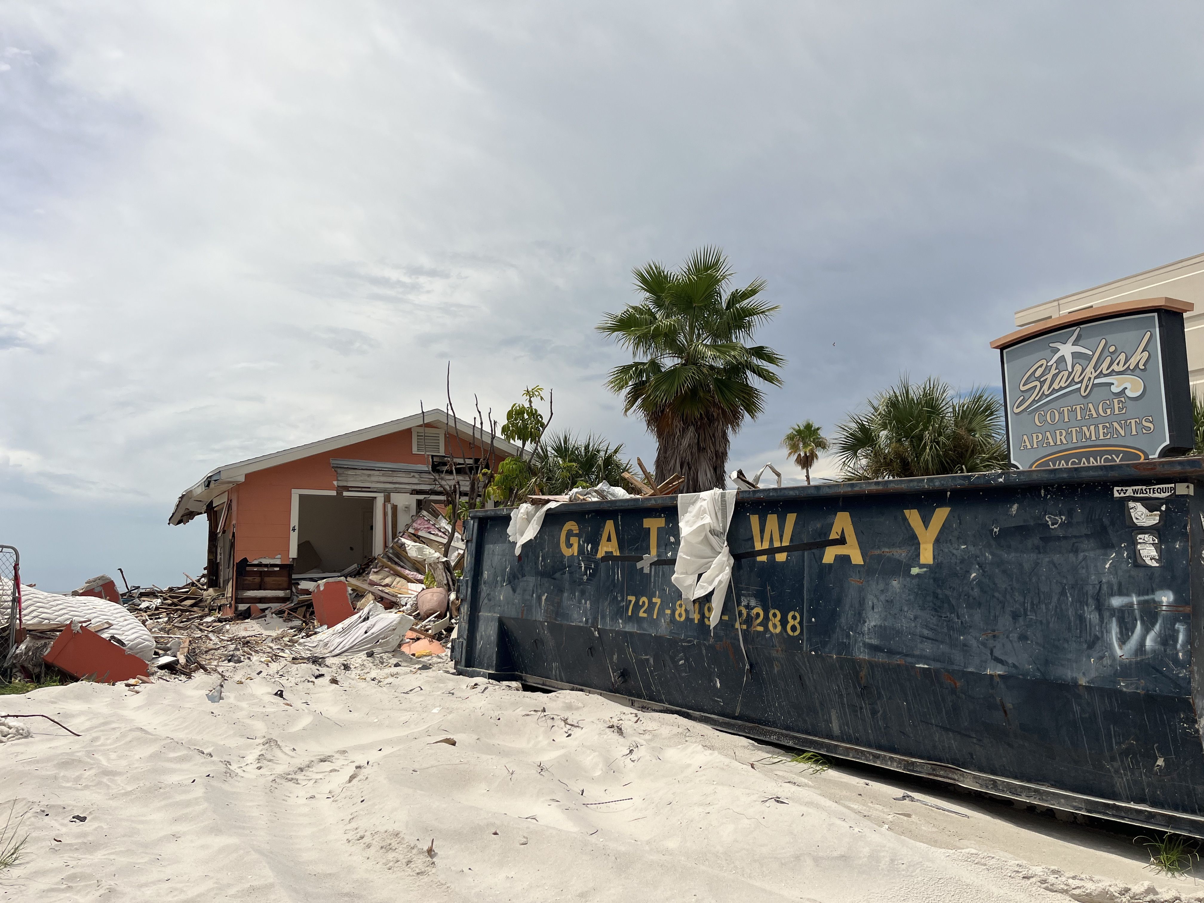 Debris and rubble from a partially collapsed orange building next to a large blue dumpster with yellow letters reading "GATEWAY" on sandy ground, under a cloudy sky near palm trees.