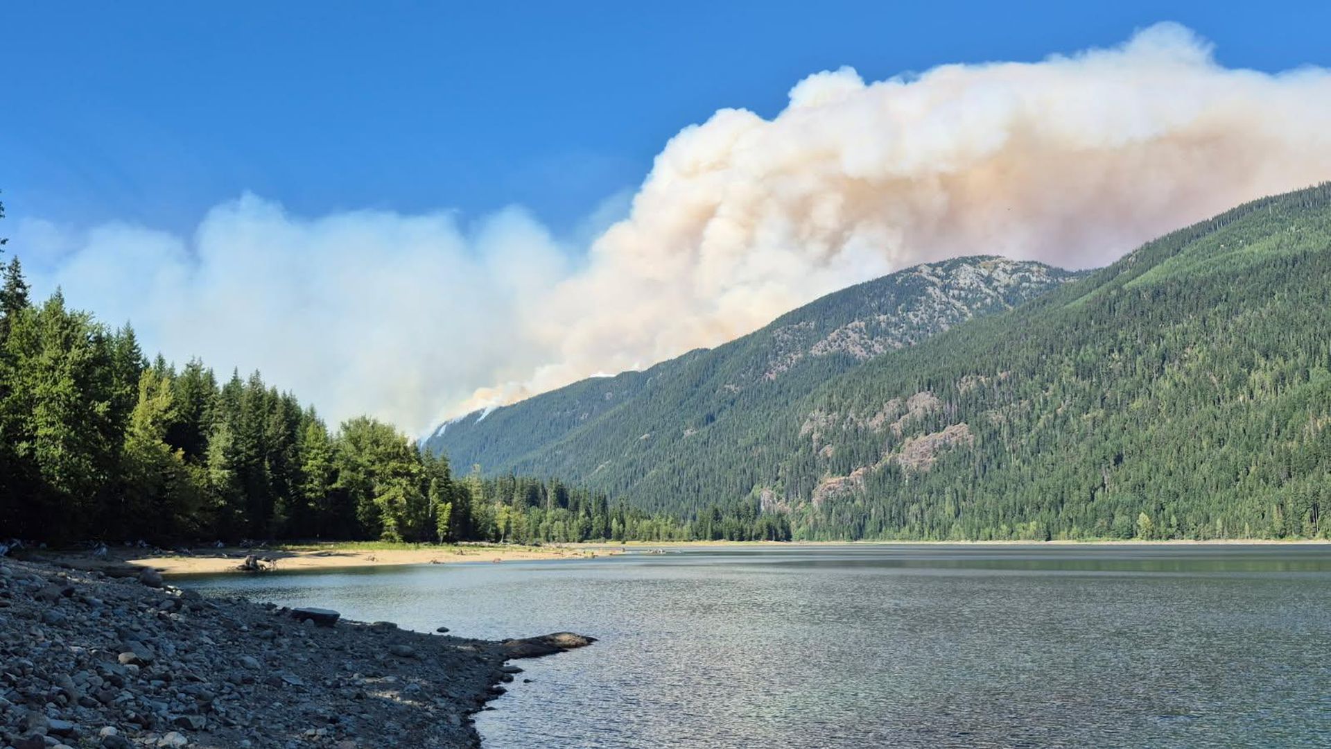Smoke rises behind a clear lakefront with rocky shore in foreground, dense evergreen forest along left and rolling forested mountains on right.