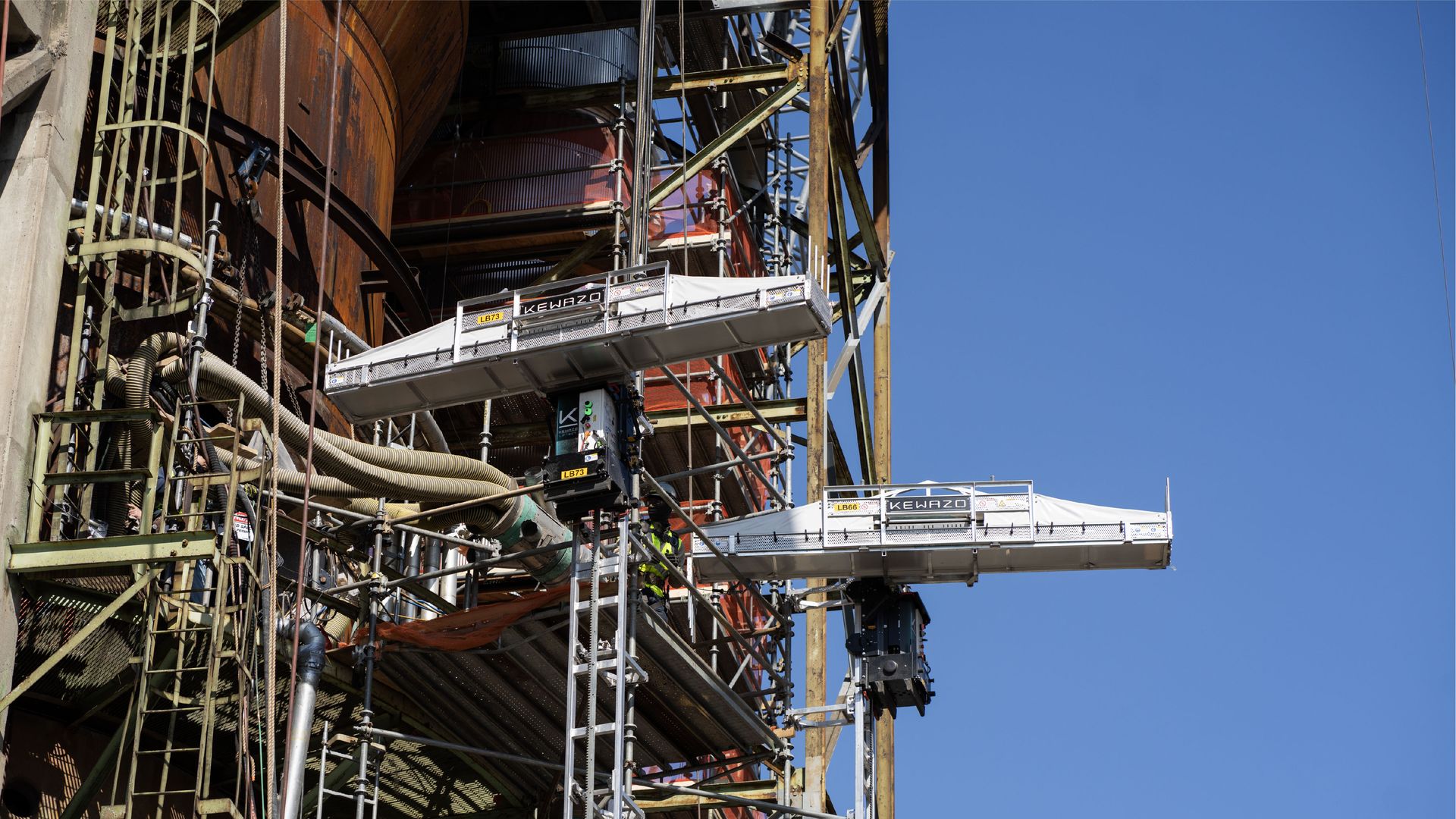 Industrial construction site with a rust-colored cylindrical structure and dense scaffolding. Two white suspended work platforms labeled KEWAZO hang above against a clear blue sky.