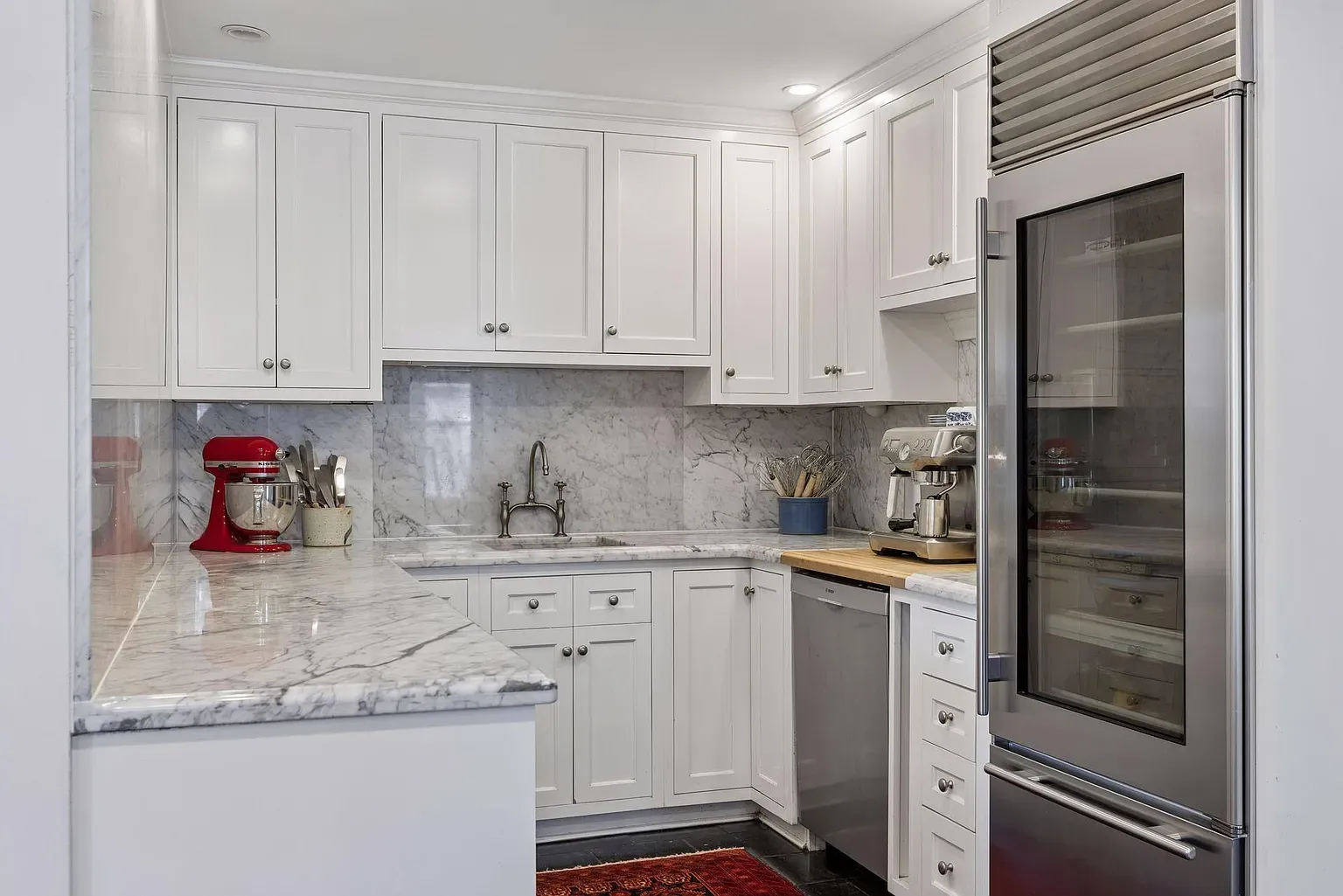 Bright white kitchen with marble countertops and backsplash, white cabinets, a tall stainless appliance with a glass door, a red stand mixer, a sink with an arched faucet, and a blue utensil jar.