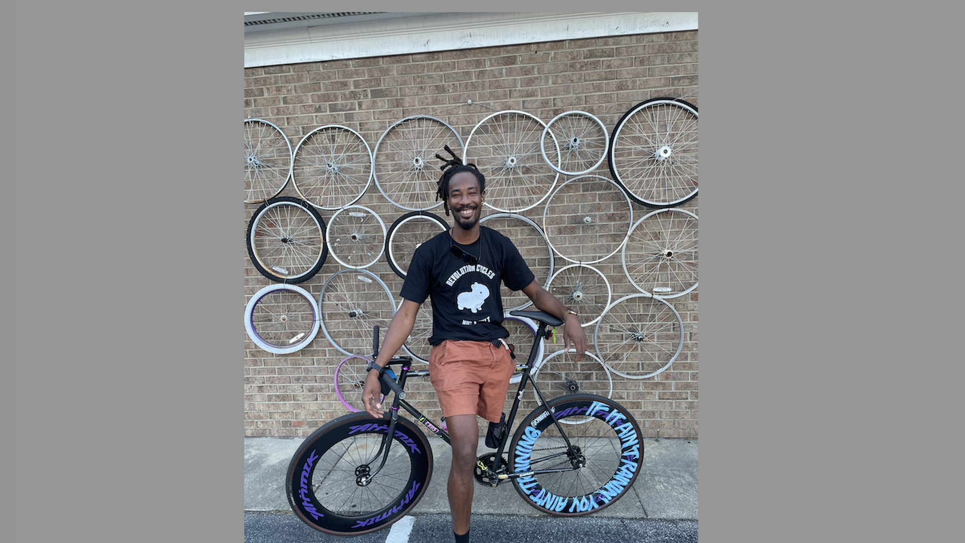 A man with one leg stands in front of a bicycle and wall with bike tire rims. 