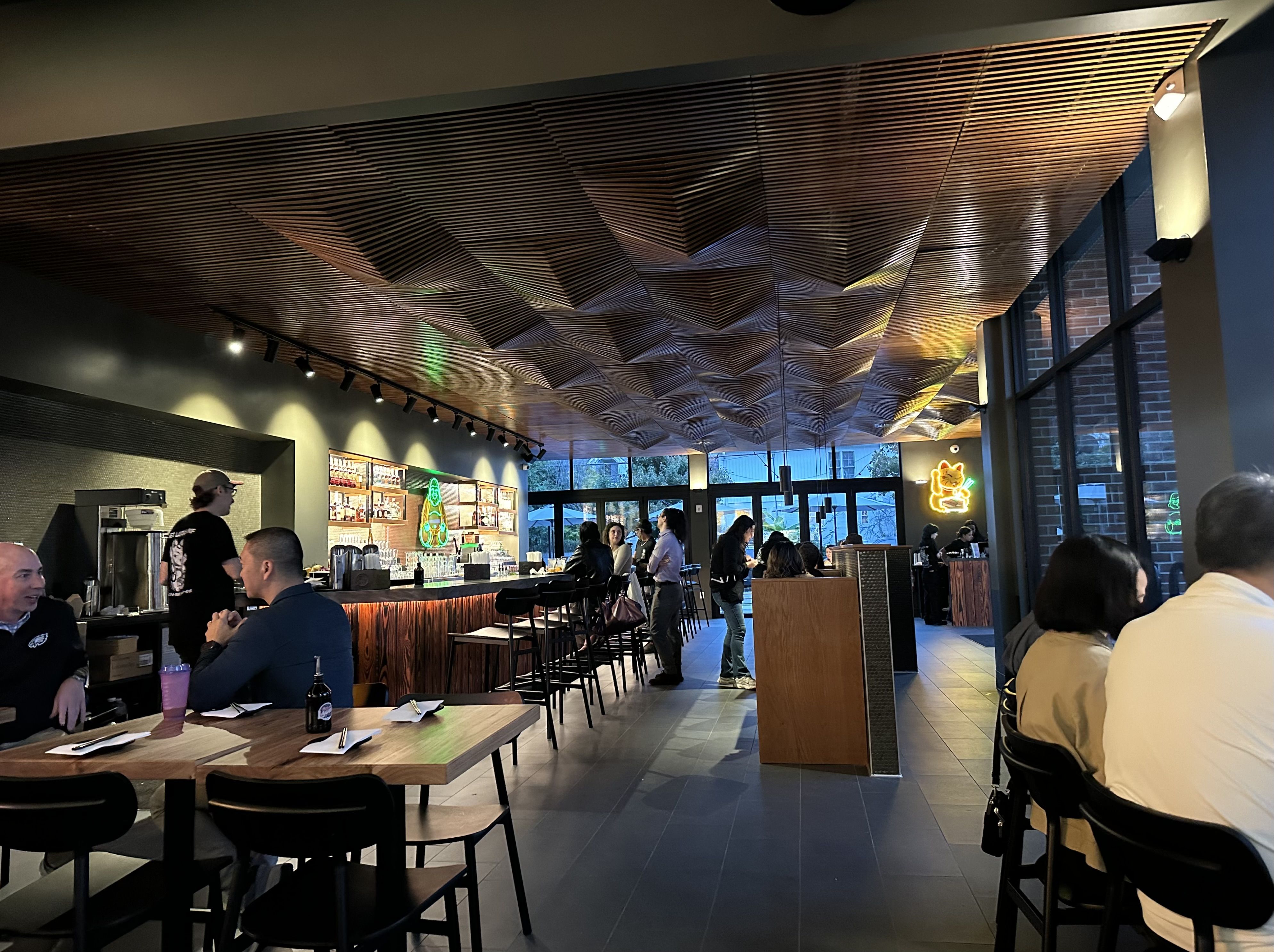 Interior of a modern cafe featuring a dramatic geometric wooden ceiling, warm pendant lighting, a long bar with stools, customers seated and chatting, neon signs near large windows and brick walls.