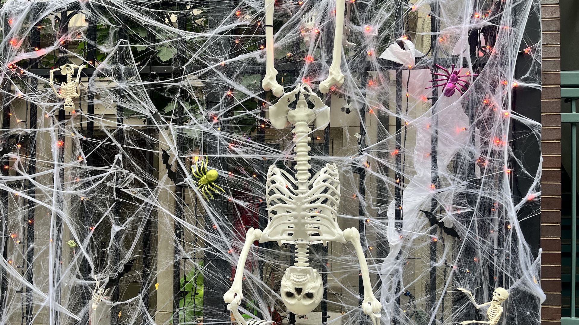 A Halloween scene with a large central skeleton and smaller skeletons tangled in white fake spiderwebs, orange LED lights along a black fence, and a purple spider in the background.