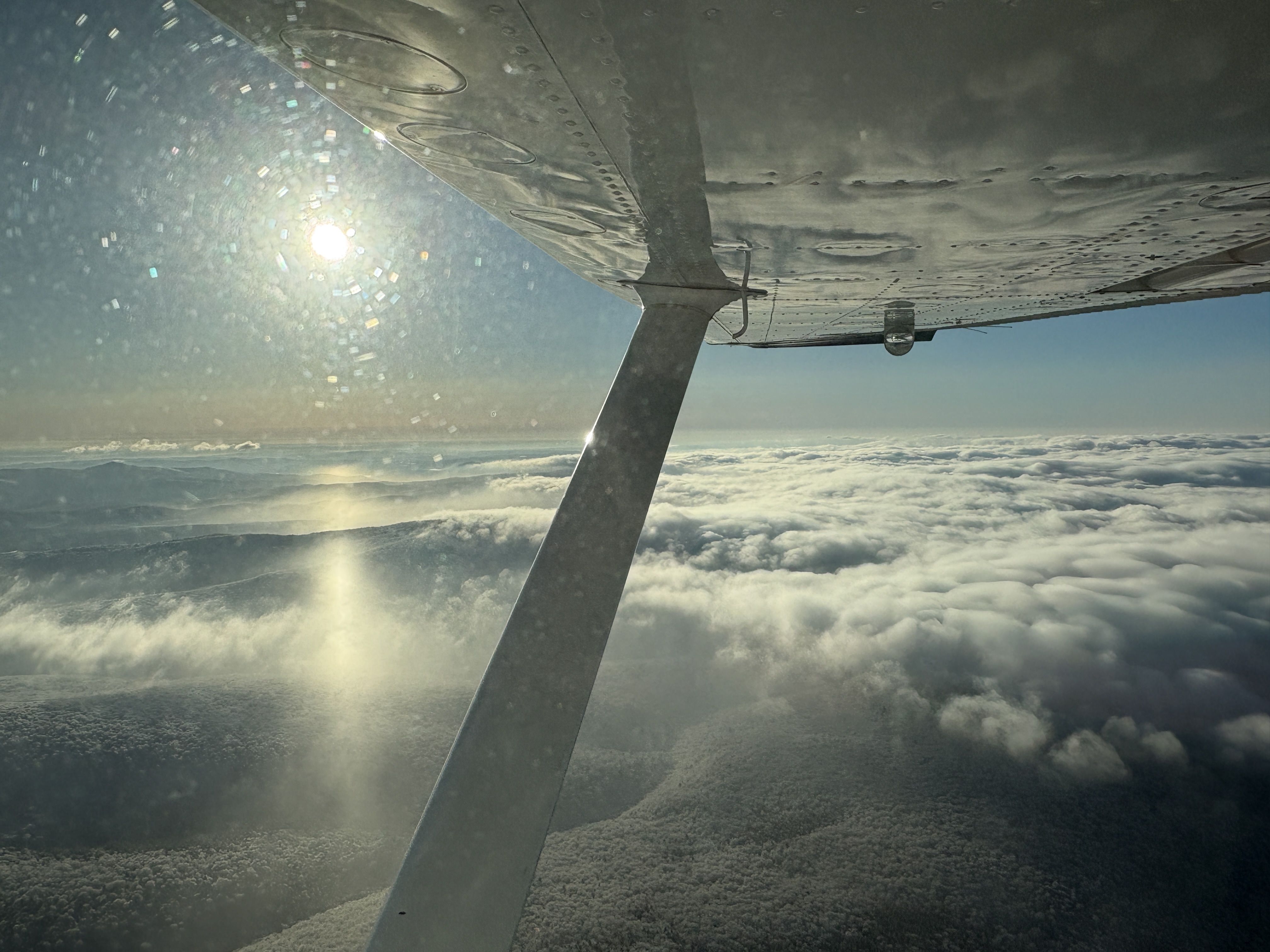 View from a small airplane window showing a metal wing with a strut, bright sun glare and window specks. Below lies a sea of fluffy clouds over distant mountains, under a blue sky.