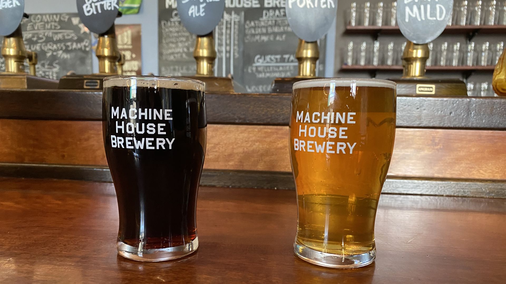 Two beers, one dark and one lighter, on a wooden bar with taps in the background. The glasses say "Machine House Brewery." 