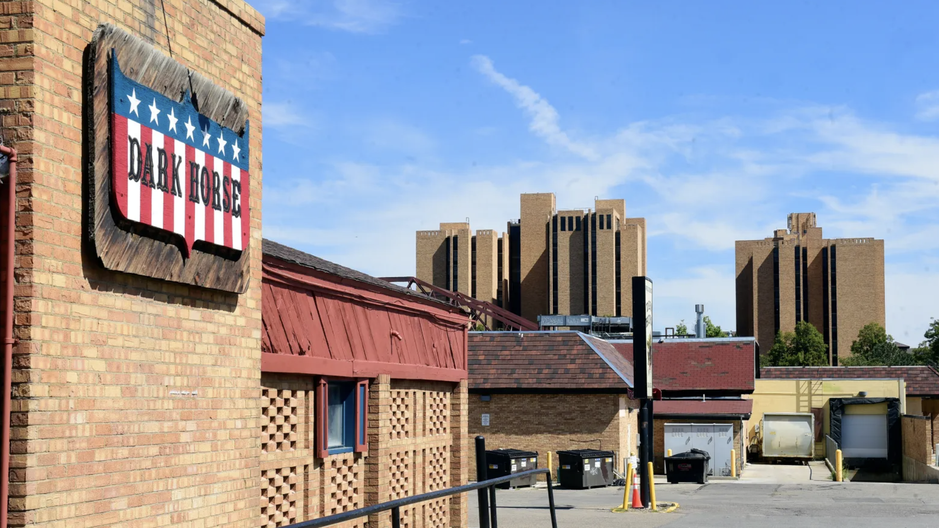 Brick building with a red, white, and blue sign reading "DARK HORSE" with stars, under a clear blue sky, parking lot with dumpsters, and large beige apartment buildings in the background.