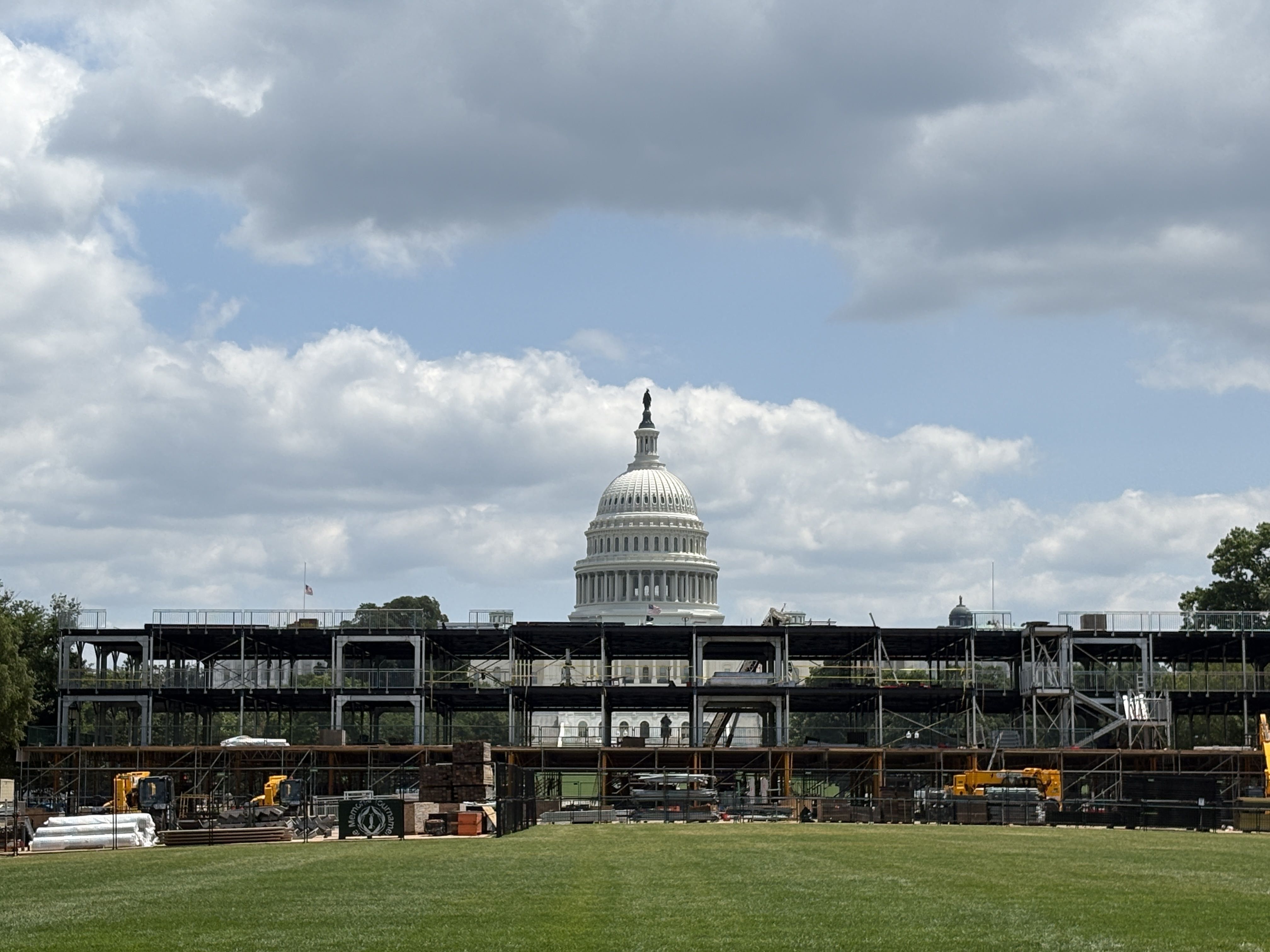 Capitol dome peeks over extensive construction scaffolding on a grassy field, with a blue sky and clouds in the background.