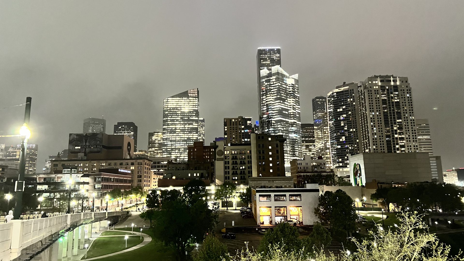 Night view of a city skyline with tall, illuminated buildings, a park with walking paths and trees in the foreground, and cloudy skies above.