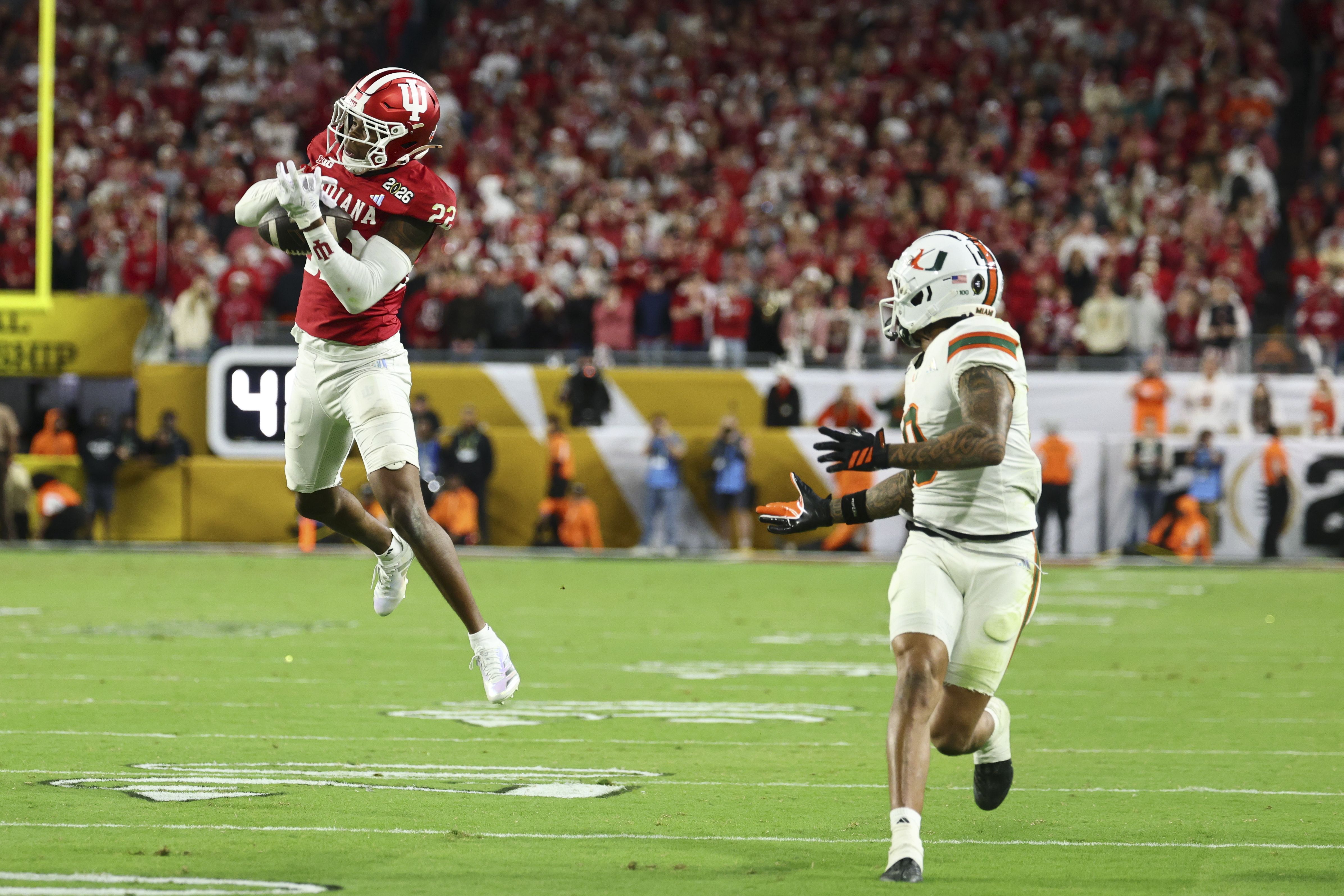 Football player in red Indiana uniform jumping to catch ball as player in white Miami uniform reaches out on green field with crowd in background.