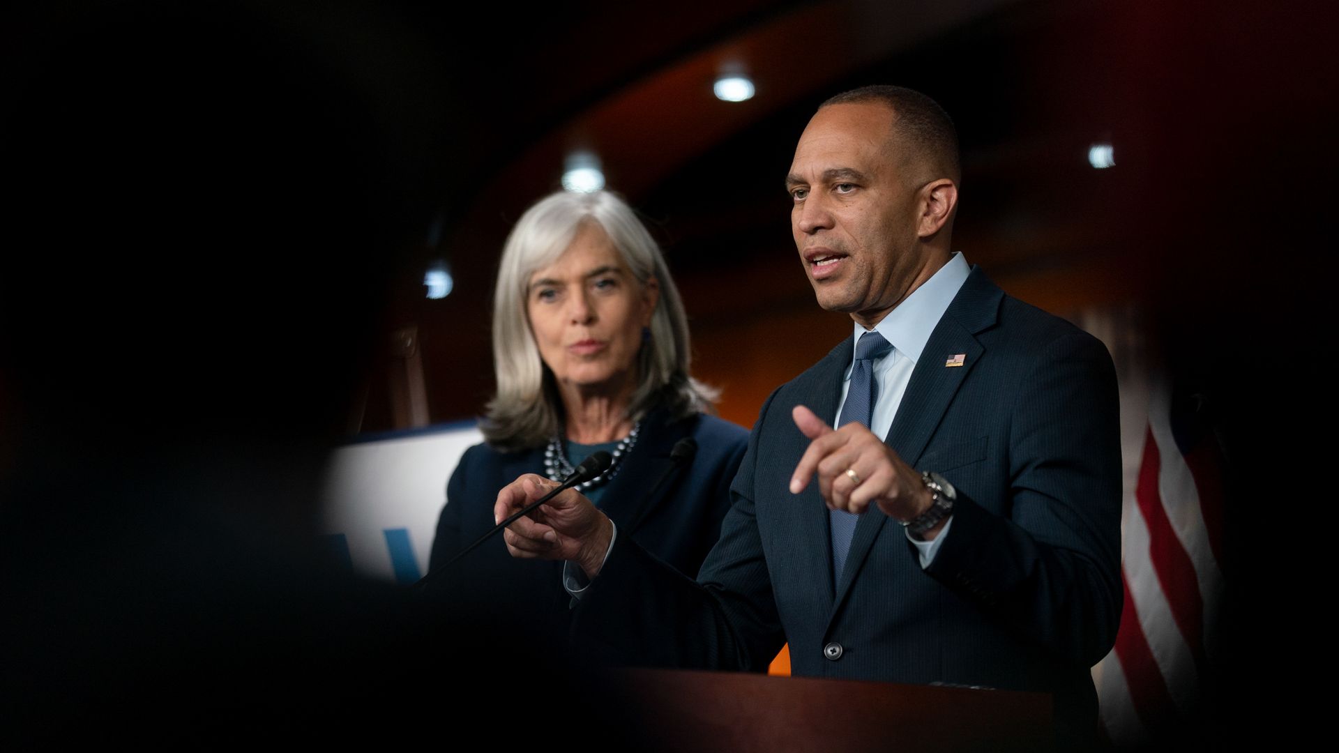 Reps. Hakeem Jeffries and Katherine Clark, both wearing  suits, are seen through a black silhouette in a brown room.