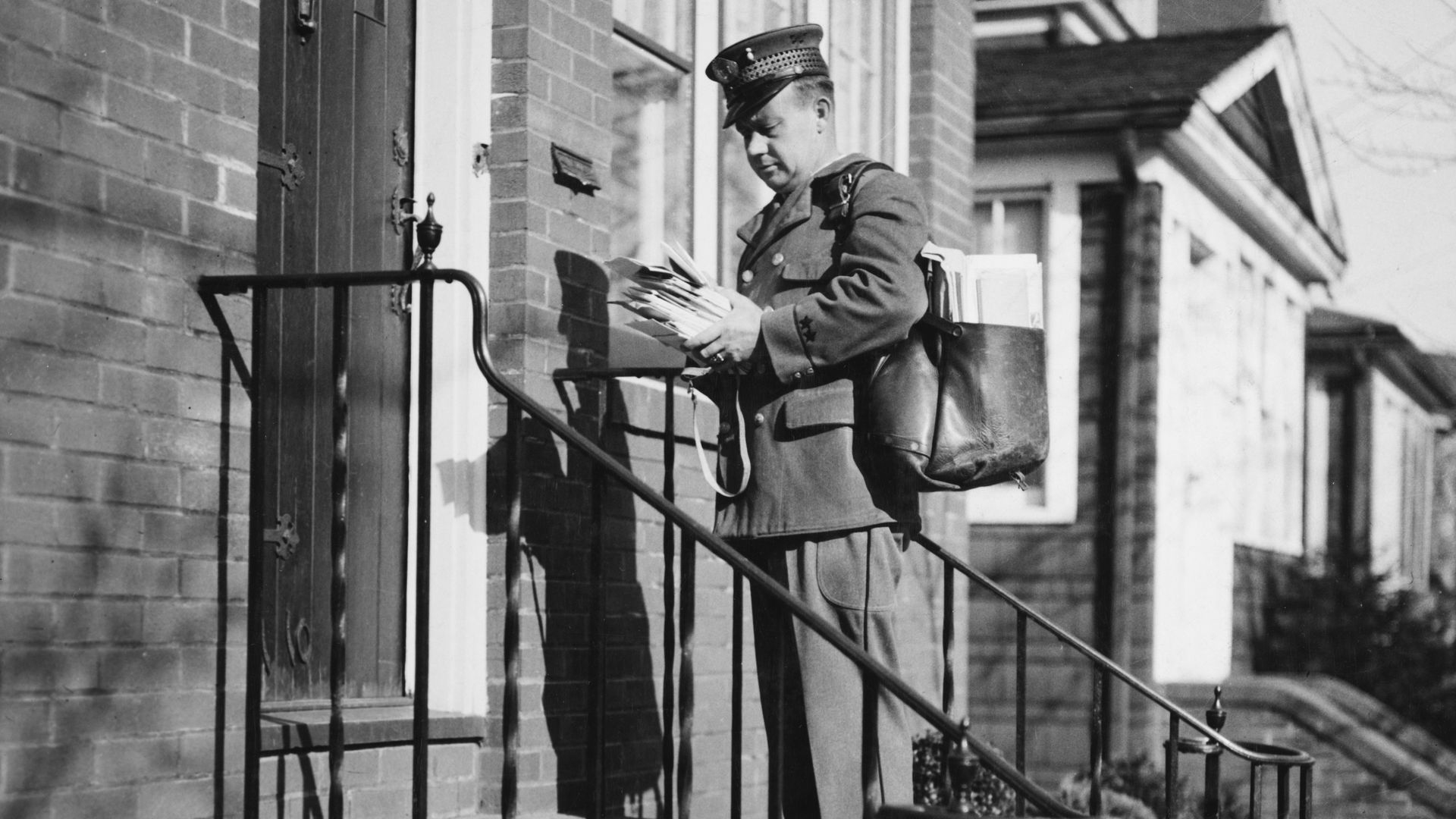 A postal carrier looks at a stack of mail on a doorstep. 