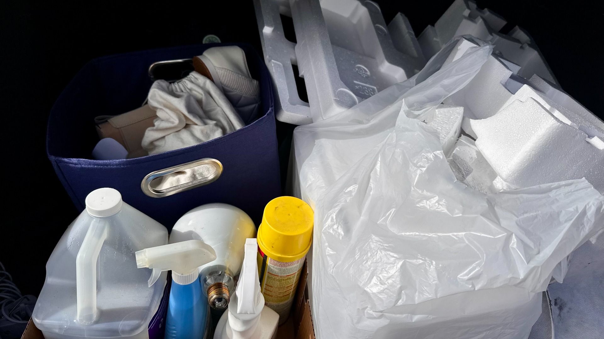A collection of recyclables including a blue storage bin with beige shoes and cloth, plastic spray bottles, a yellow canister, a large white plastic bag, and white styrofoam pieces.
