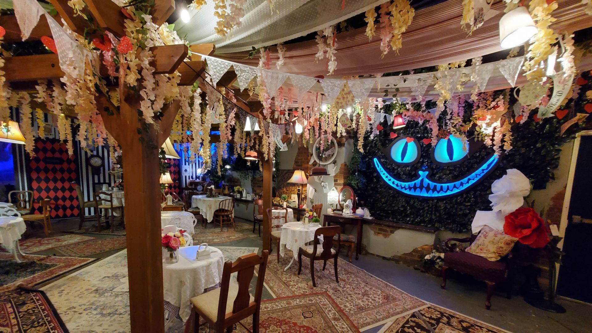 An Alice in Wonderland themed room with flowers hanging from the ceiling, tea sets on round tables and a light-up Cheshire Cat grin built into the wall